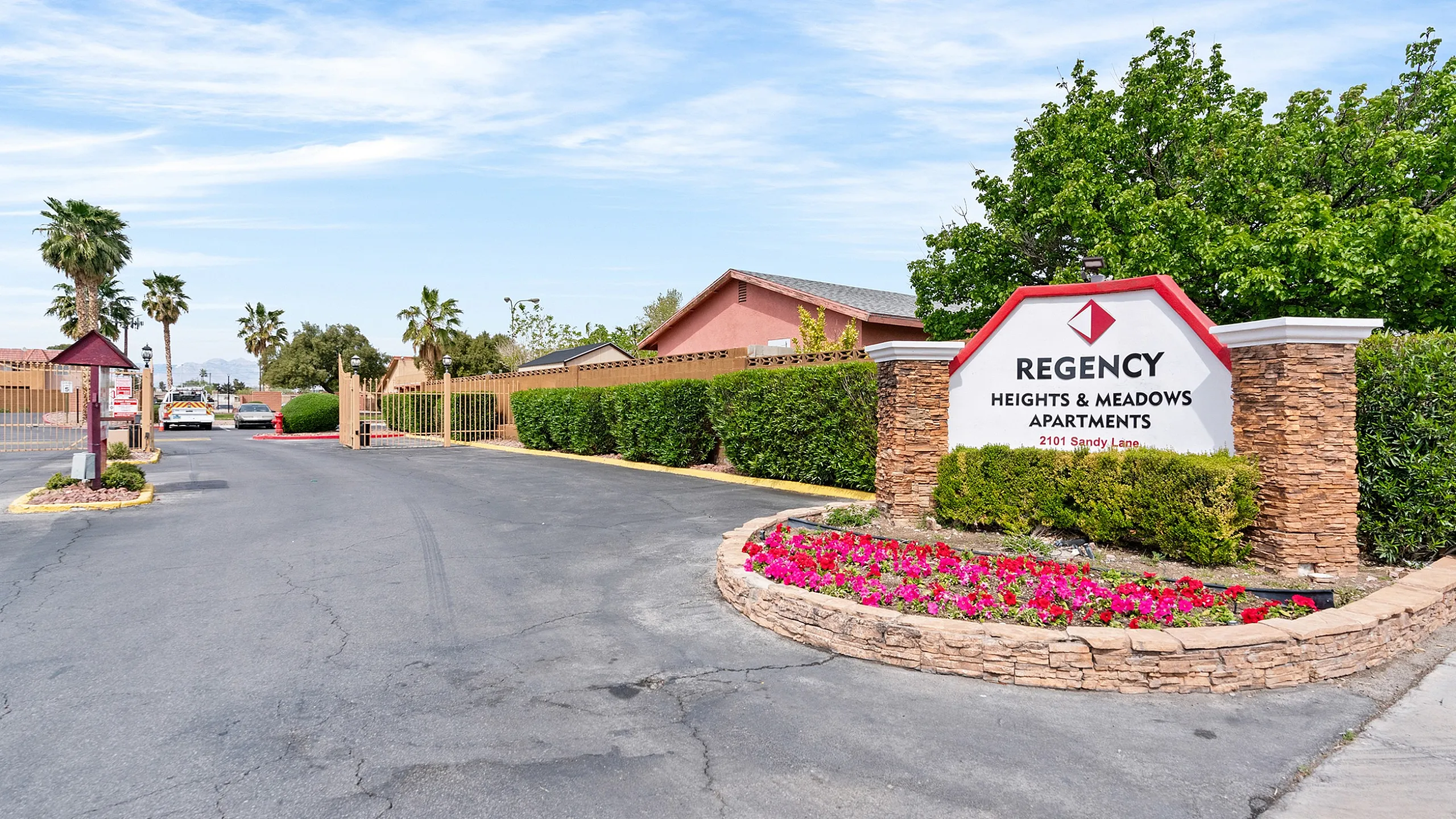 Inviting entrance to Regency Heights & Meadows Apartments, featuring lush landscaping and vibrant flowers under a clear blue sky.
