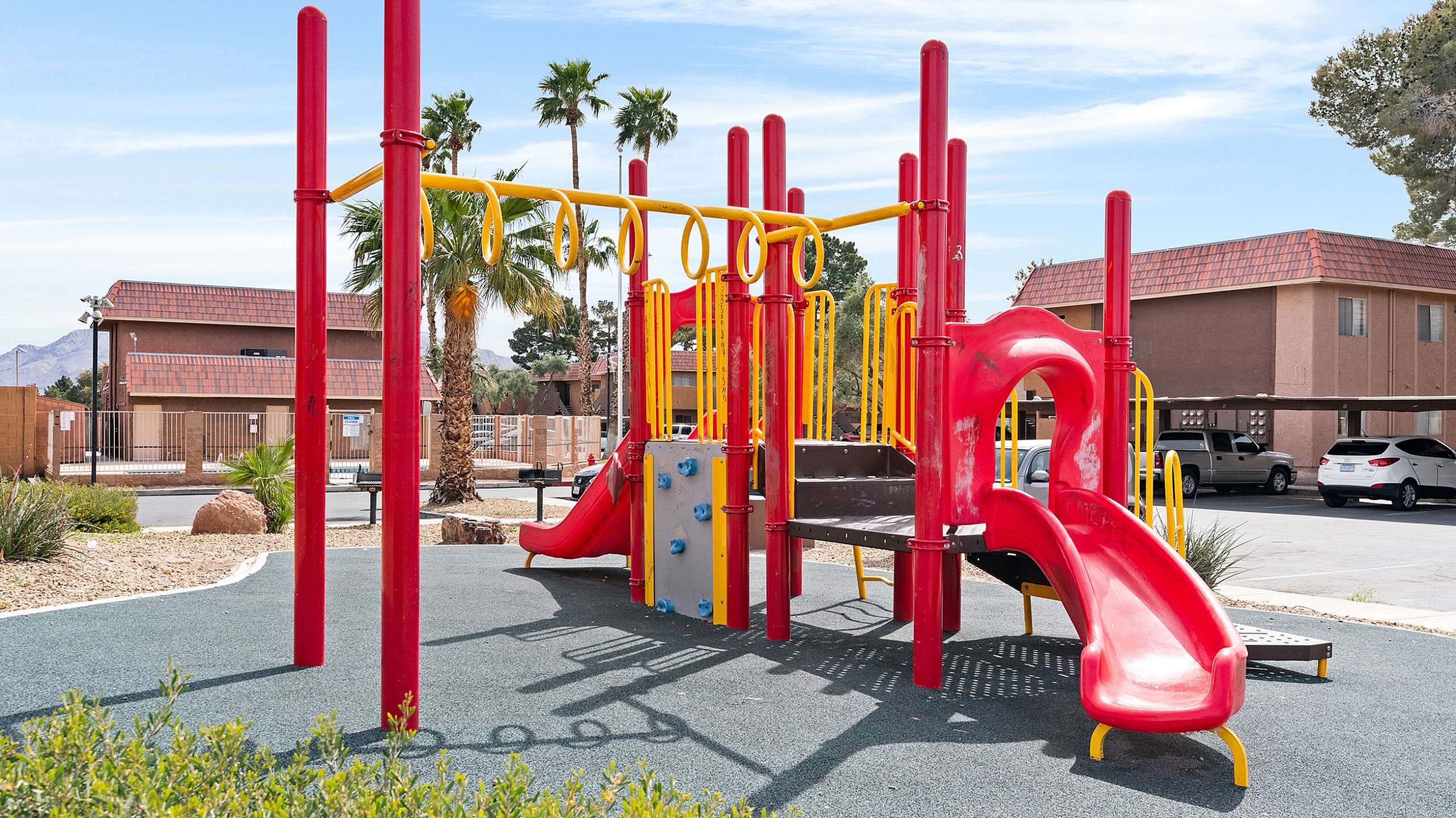 Vibrant playground with red and yellow equipment, featuring slides and climbing structures, surrounded by palm trees and clear blue skies.