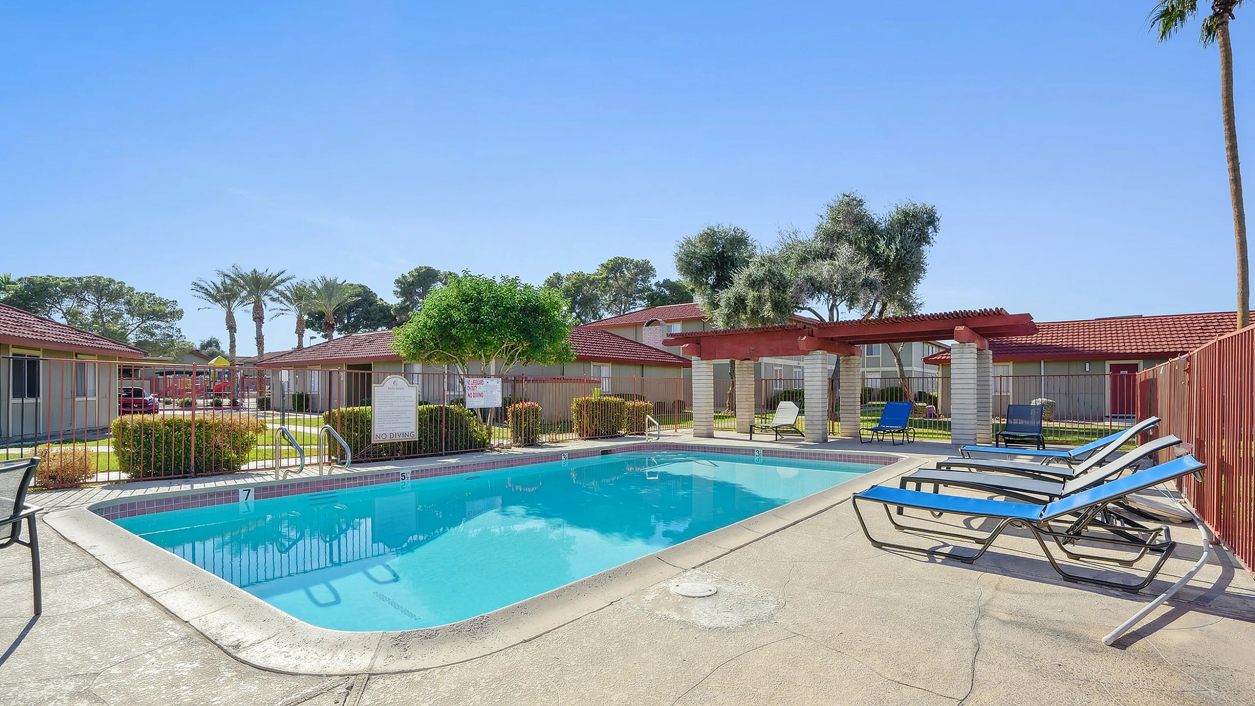 Inviting apartment complex pool area with sun loungers, surrounded by lush greenery and vibrant red-roofed buildings, perfect for relaxation.