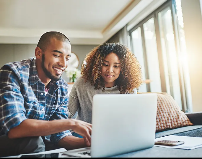 Cheerful couple working on a laptop in a bright, modern living space with large windows and cozy decor.