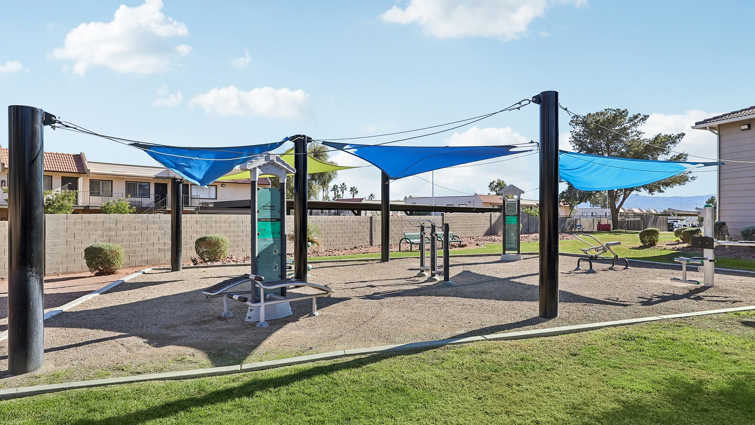 Outdoor fitness area with modern exercise equipment and vibrant sun sails, surrounded by lush greenery and a sunny sky.