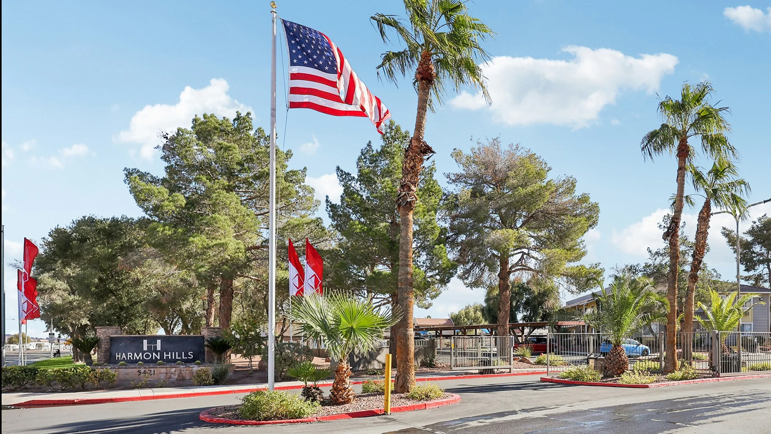 Inviting entrance of Harmon Hills with lush palm trees, vibrant flags waving under a clear blue sky, and a welcoming communal atmosphere.