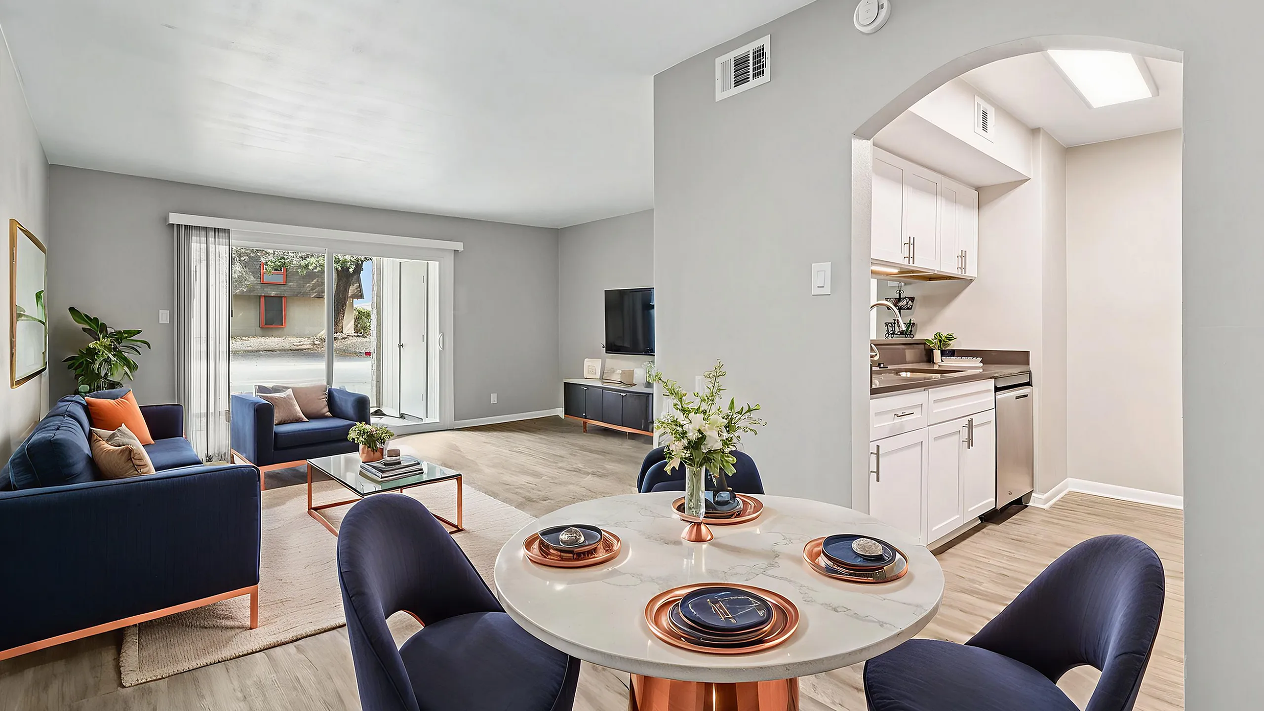 Beautifully staged apartment with elegant navy blue seating, modern dining area, and sleek kitchen. Bright living room features stylish decor and opens to a serene outdoor view.