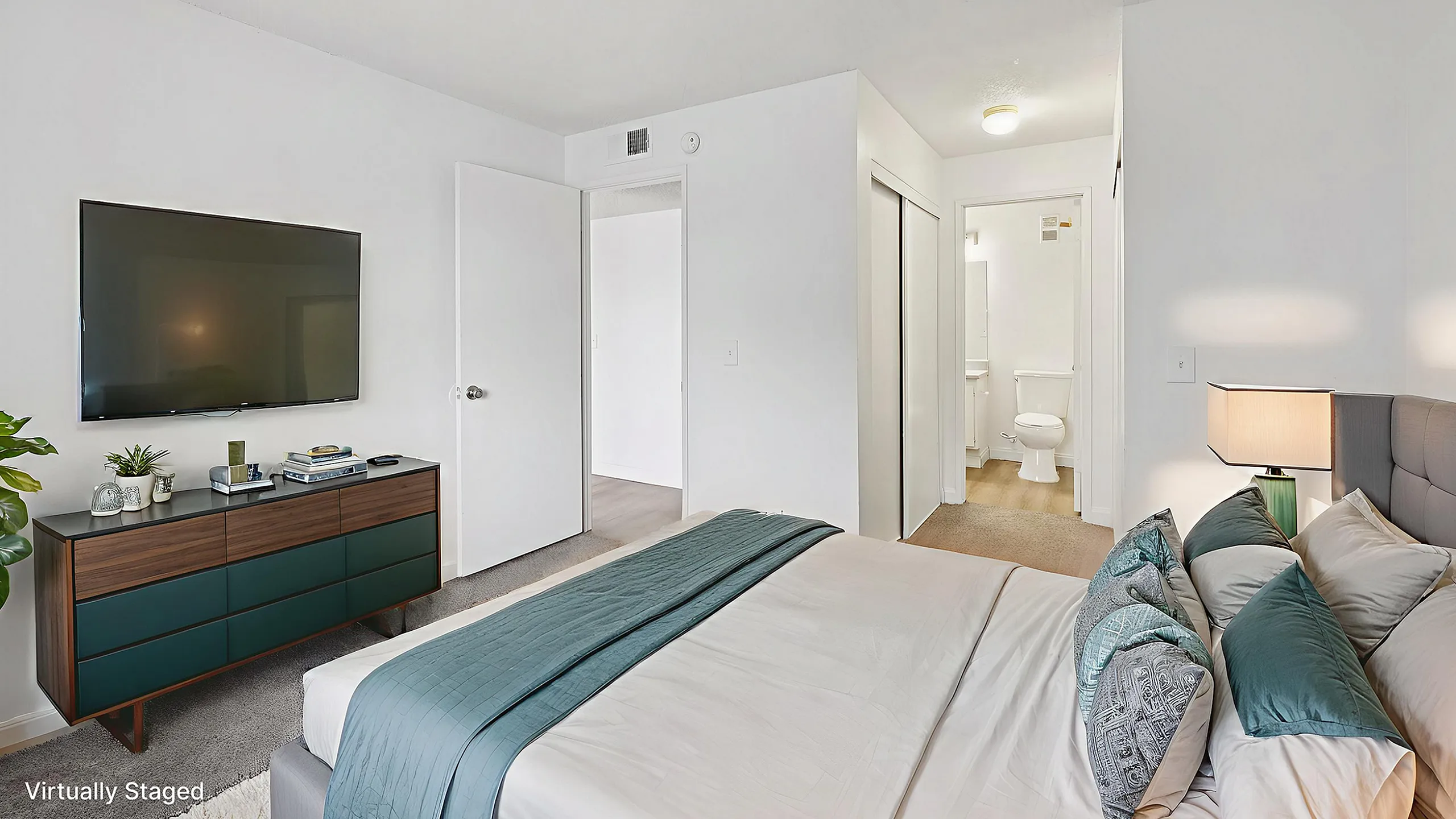 Inviting bedroom with sleek furnishings, featuring a flat-screen TV, cozy bed with plush pillows, and a modern dresser, leading into a bright ensuite bathroom.