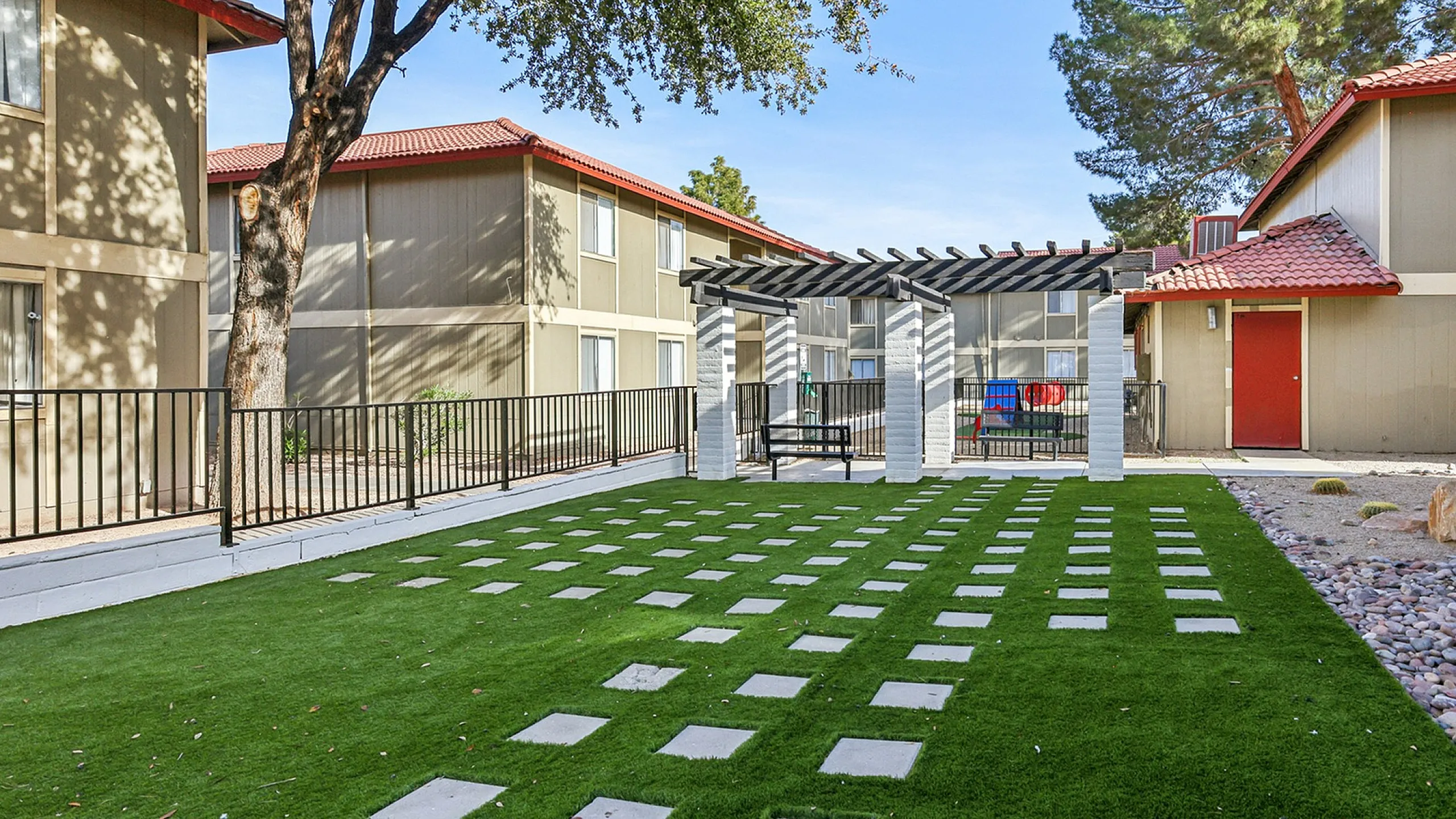 Charming apartment courtyard with lush green lawn and shaded seating area, surrounded by modern buildings with red-tiled roofs.