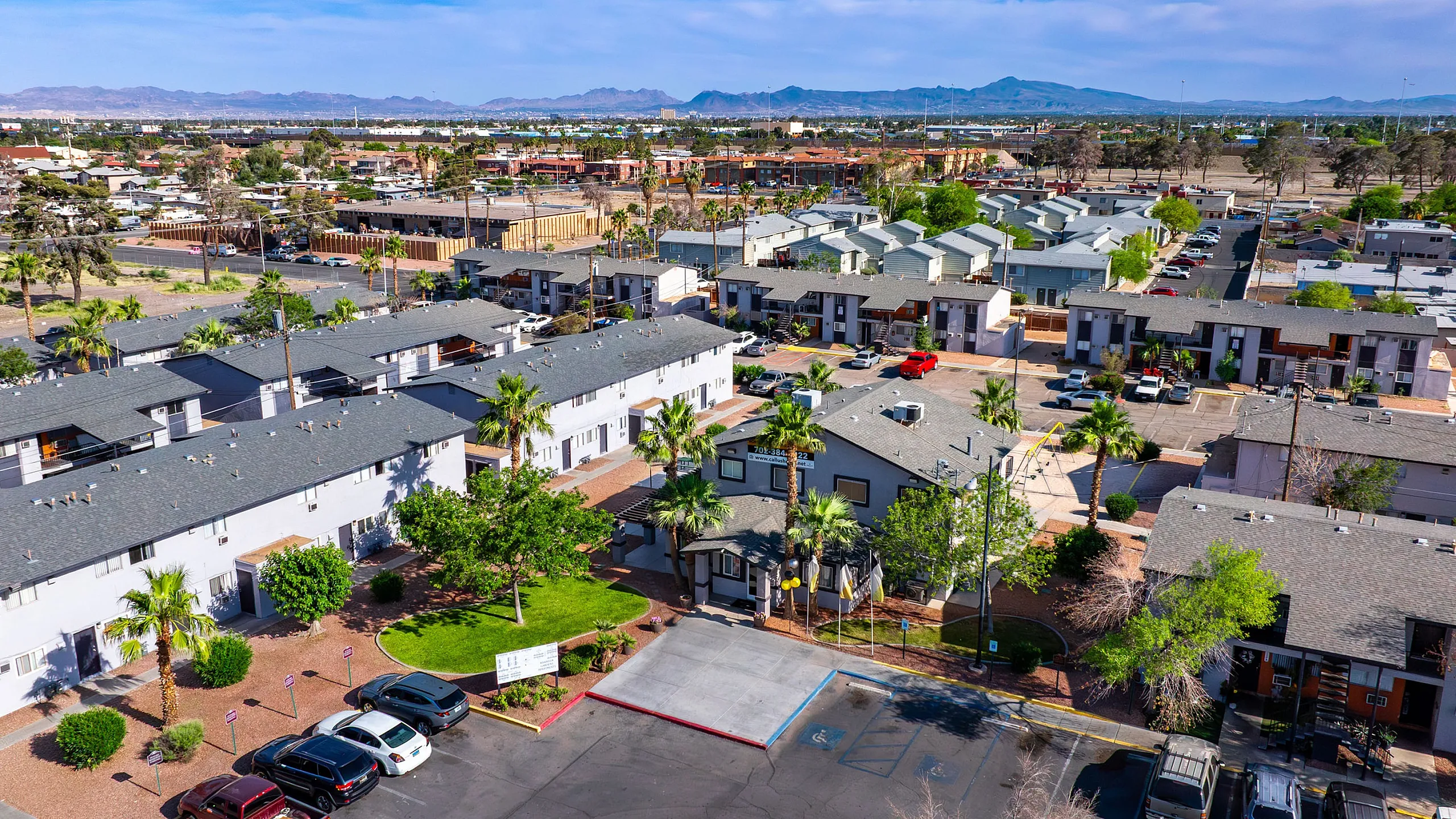 Aerial view of a charming residential community with palm trees, lush greenery, and ample parking, set against a scenic mountainous backdrop.