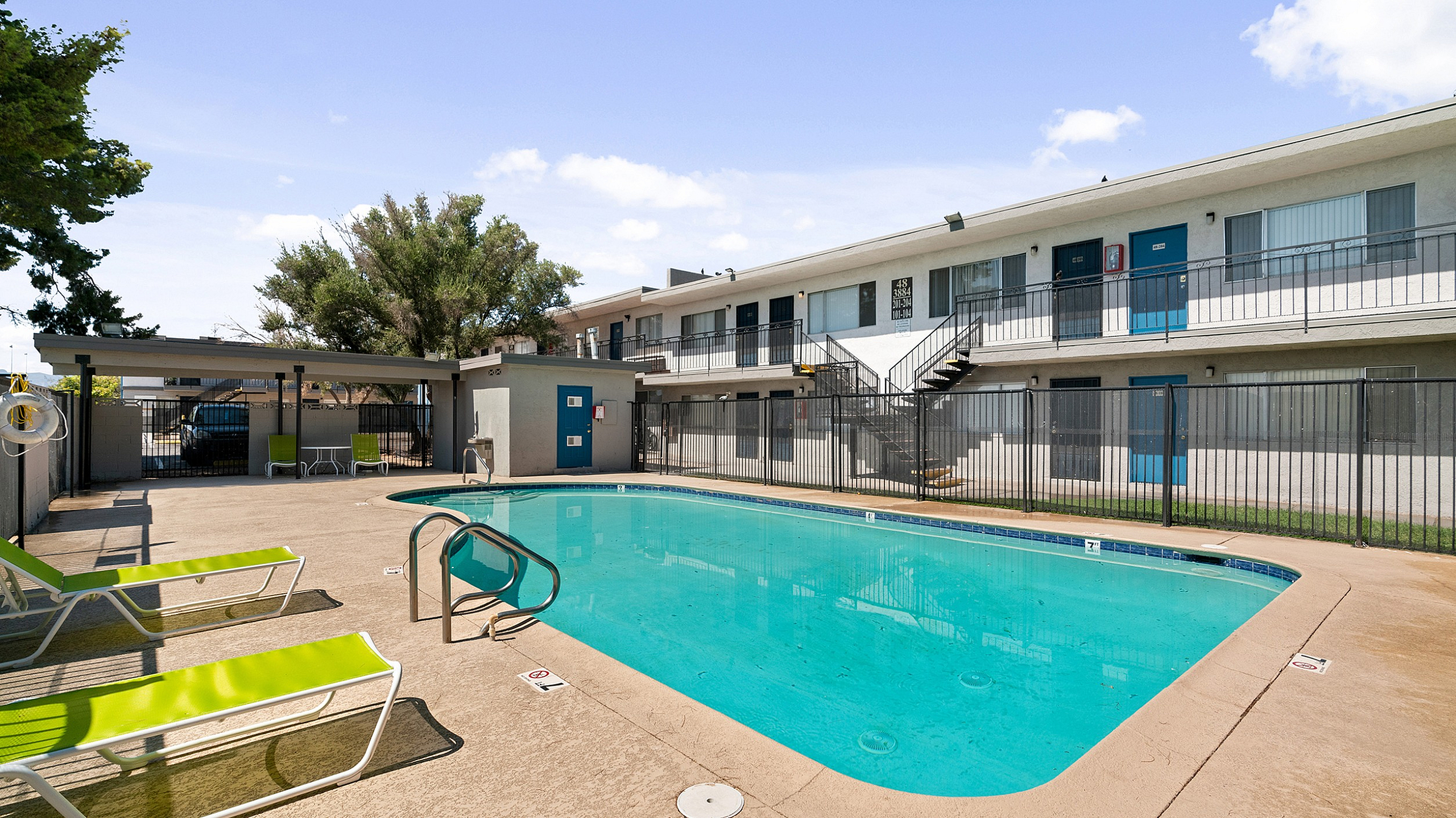Modern apartment complex featuring a sparkling swimming pool with vibrant green lounge chairs, surrounded by sleek architecture and lush trees.