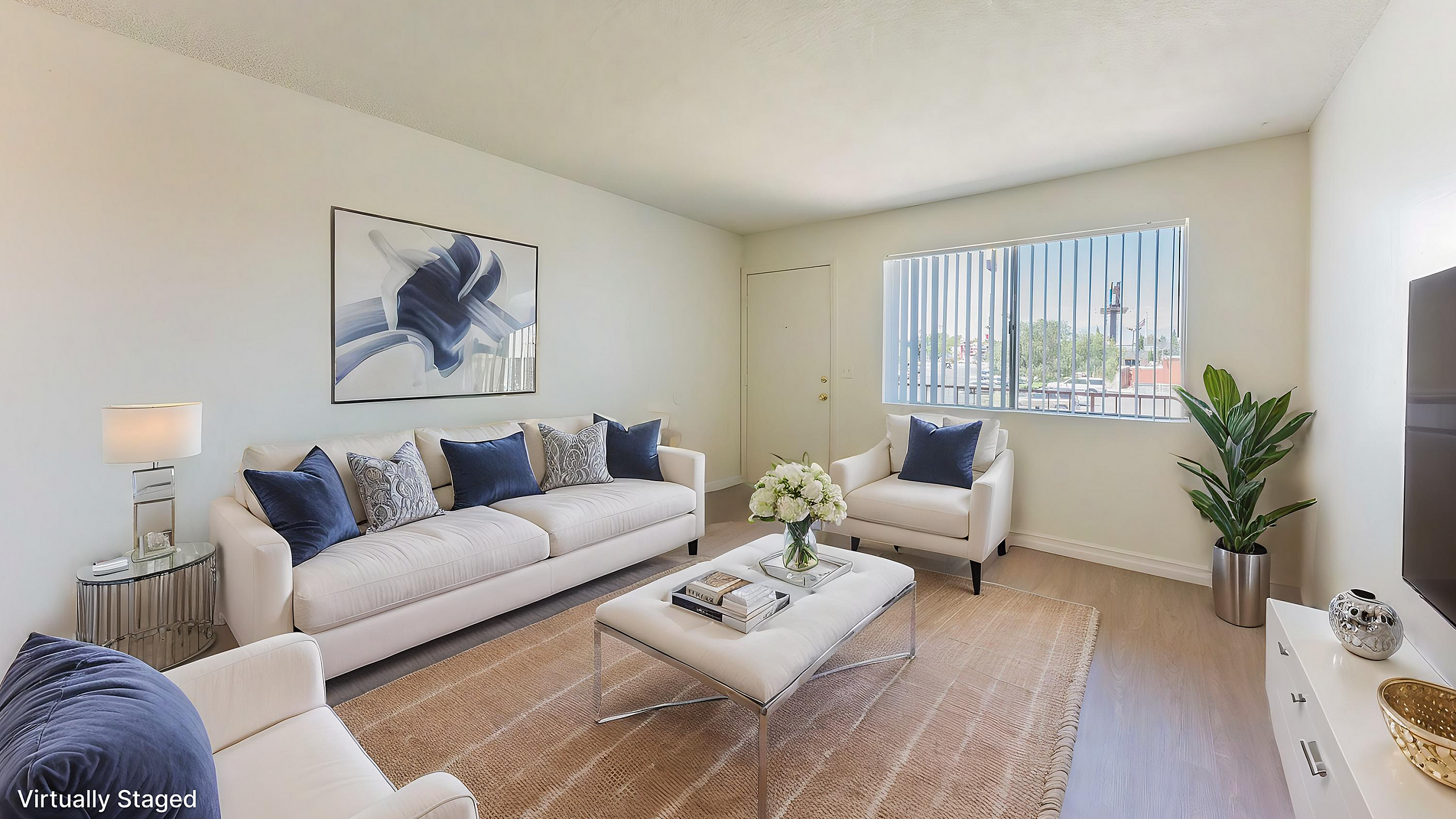 Bright living room with modern furniture, cozy white couches with blue cushions, a stylish coffee table, and a large window offering natural light. Virtually staged for a welcoming ambiance.