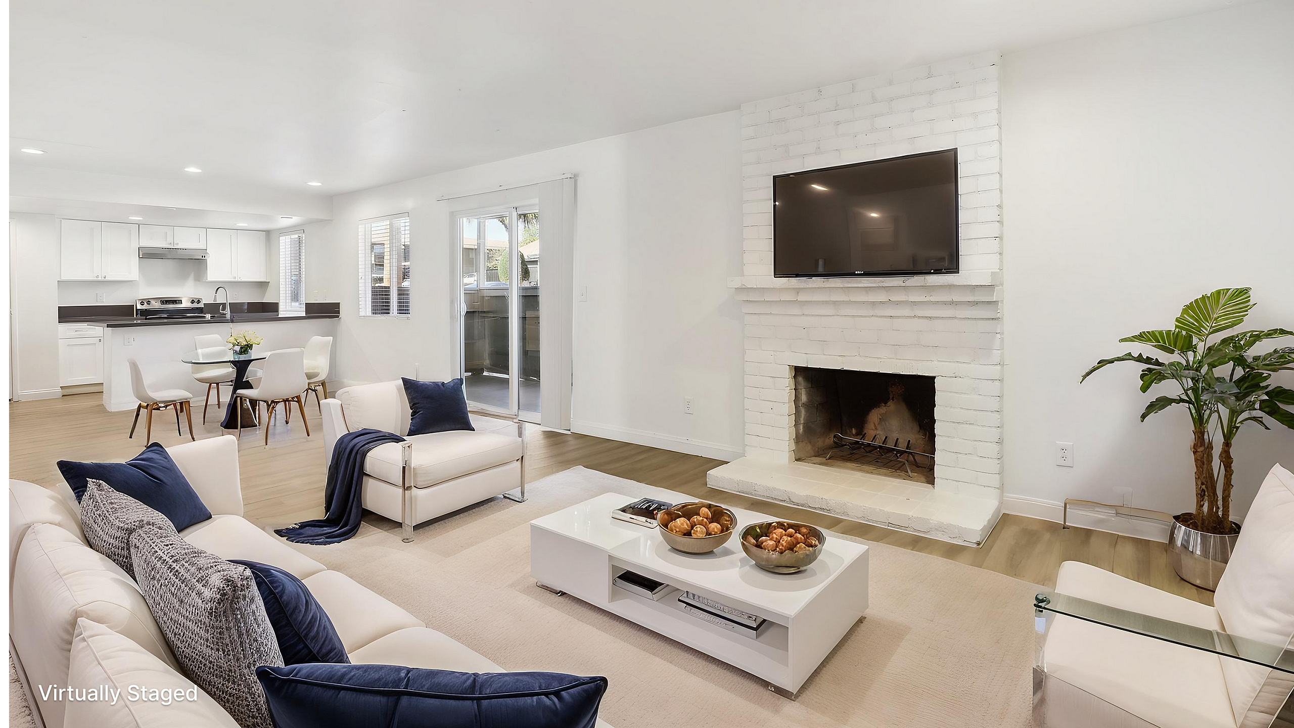 Bright and modern open-concept living room with a cozy fireplace, elegant white furnishings, and a sleek kitchen in the background.