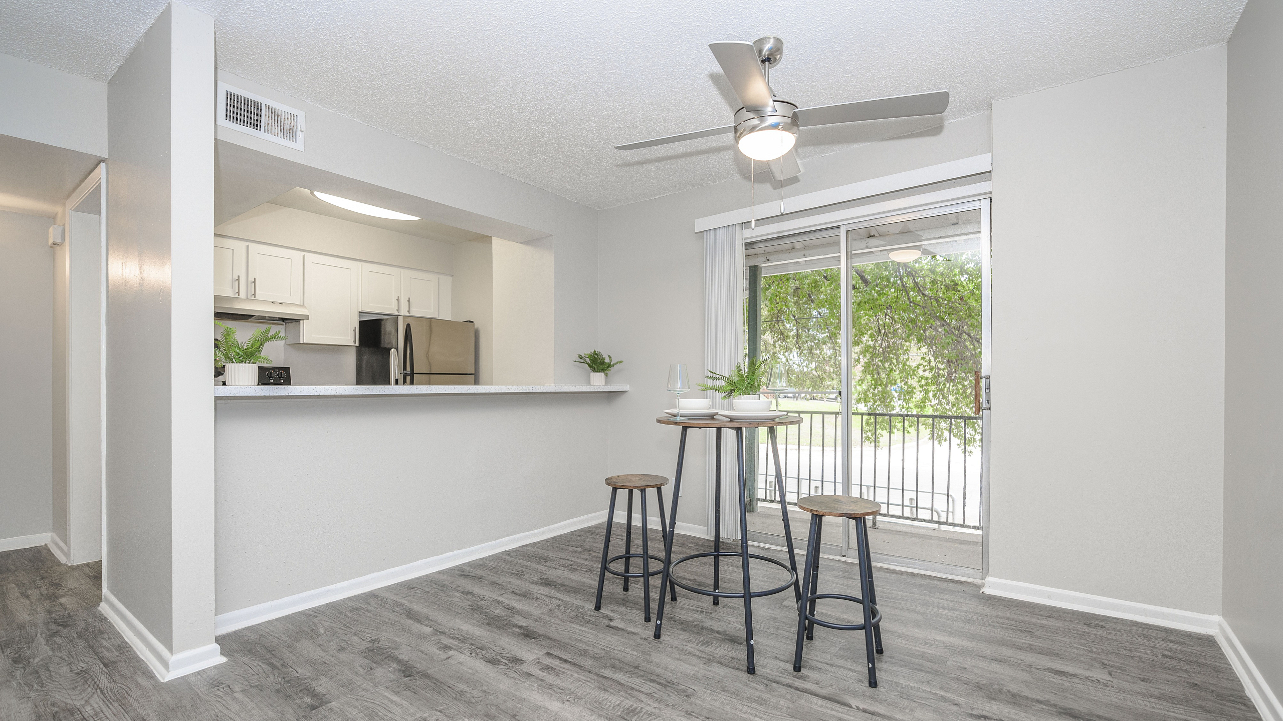 Bright and airy dining area with modern bar stools and stylish ceiling fan, overlooking a lush green view through sliding glass doors.