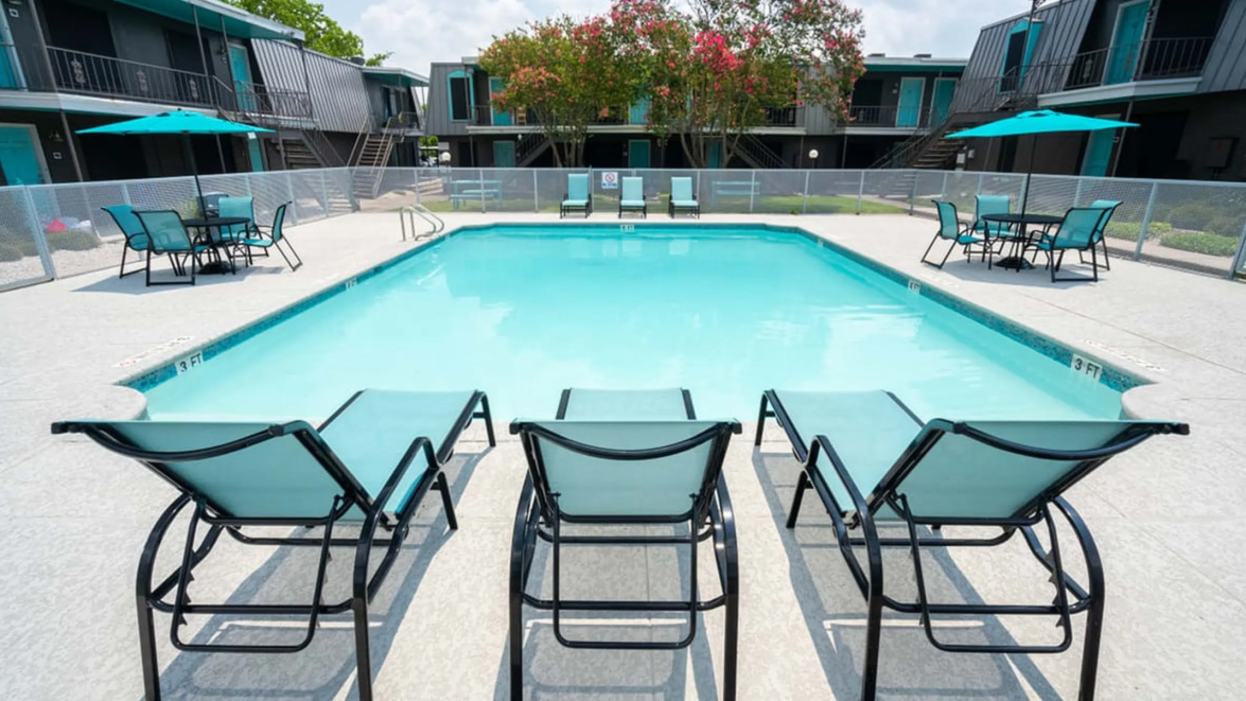Inviting apartment pool area with stylish lounge chairs and vibrant umbrellas, surrounded by modern buildings and lush greenery.