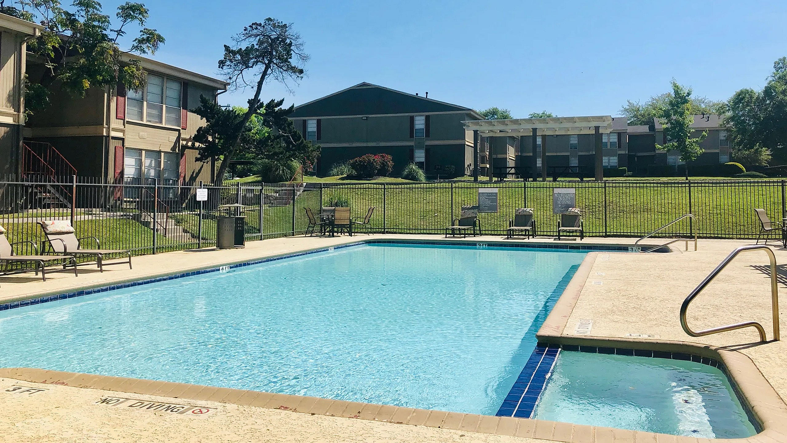 Inviting apartment complex with a sparkling community pool, surrounded by lush greenery and sun loungers under clear blue skies.