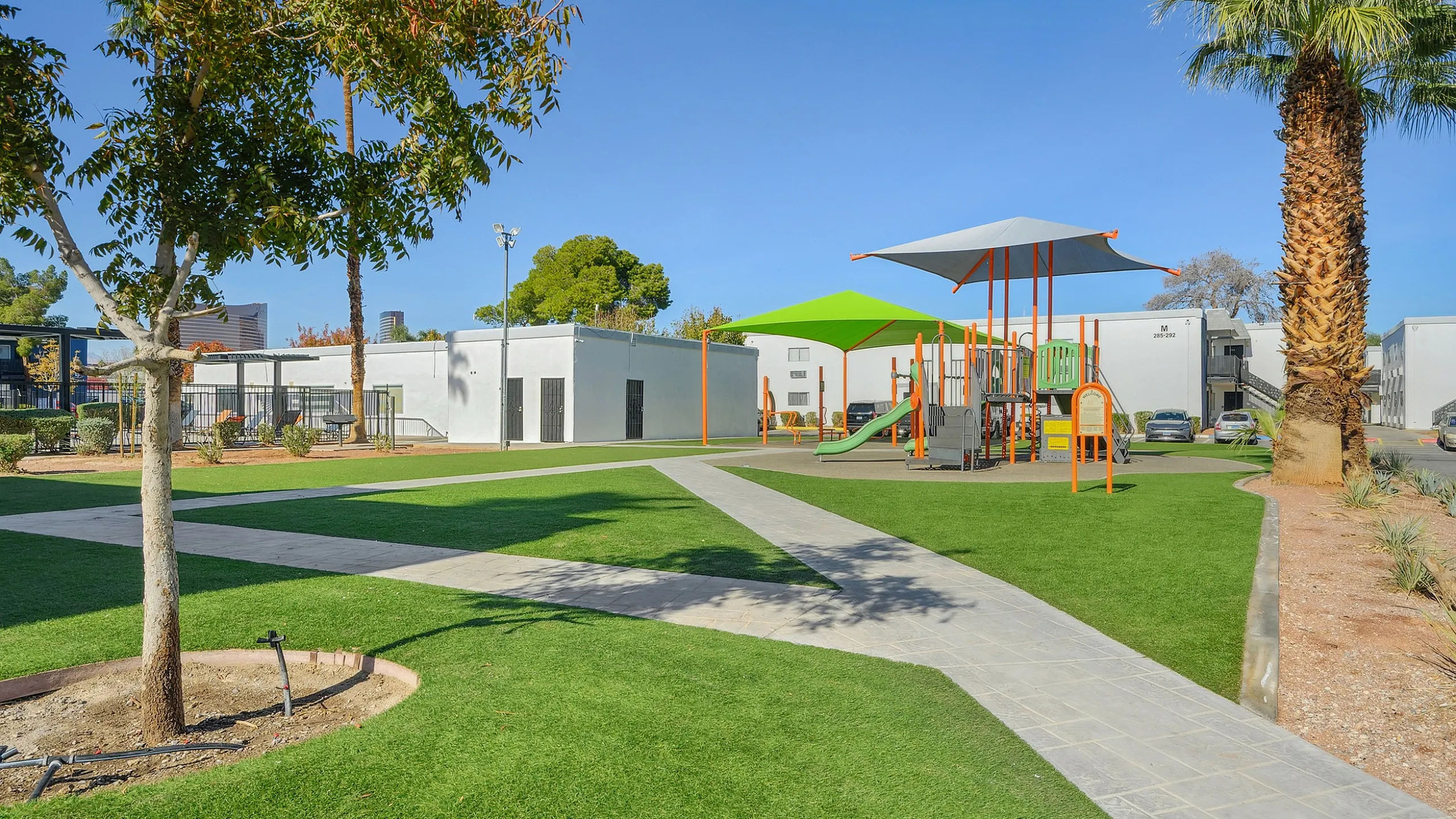 Vibrant community park featuring a modern playground with green canopies, lush grass areas, and surrounding white residential buildings under a clear blue sky.
