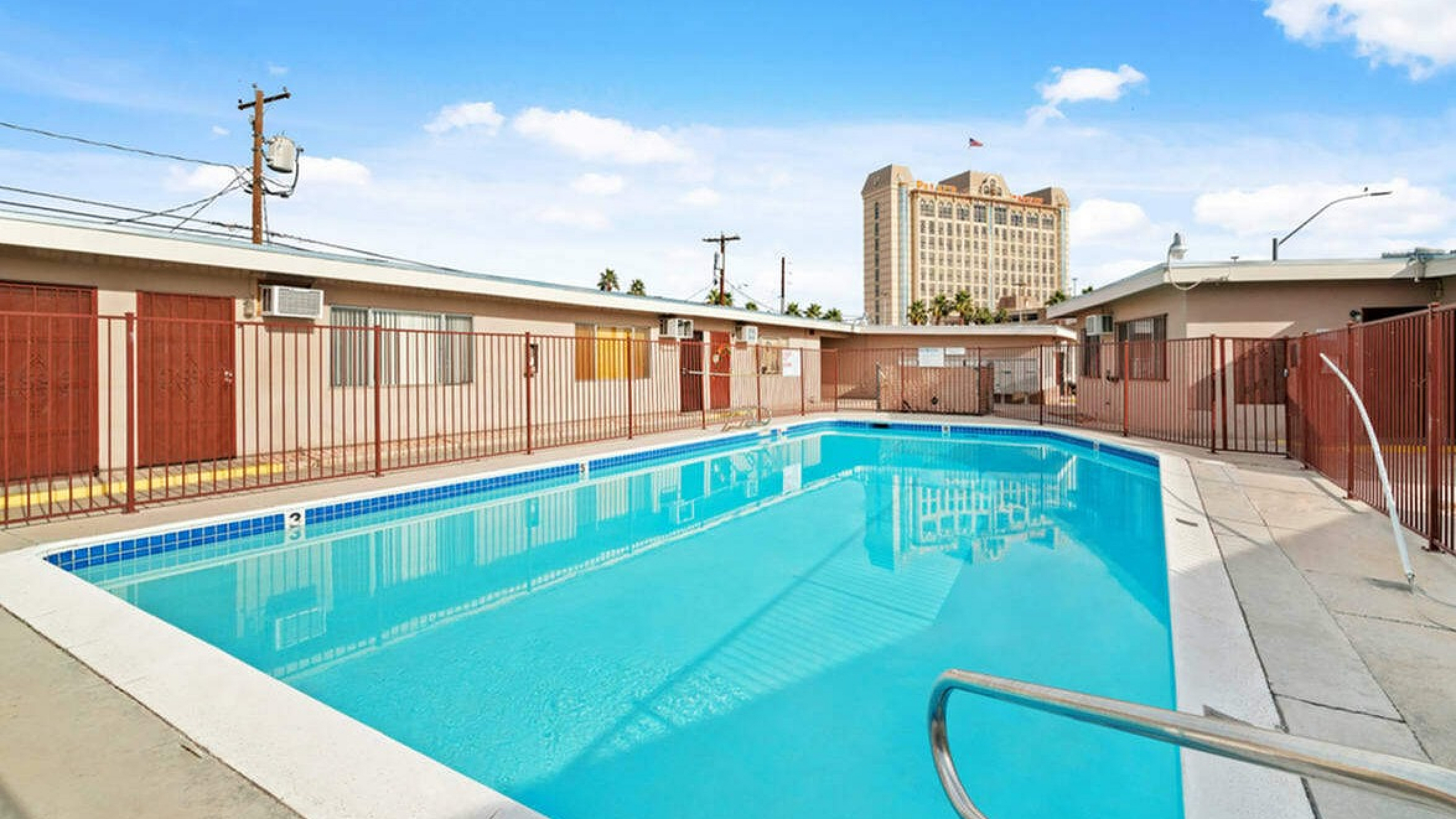 Inviting outdoor swimming pool area with vibrant blue waters surrounded by a secure fence, featuring nearby cozy apartments and a stunning building in the background under a clear blue sky.