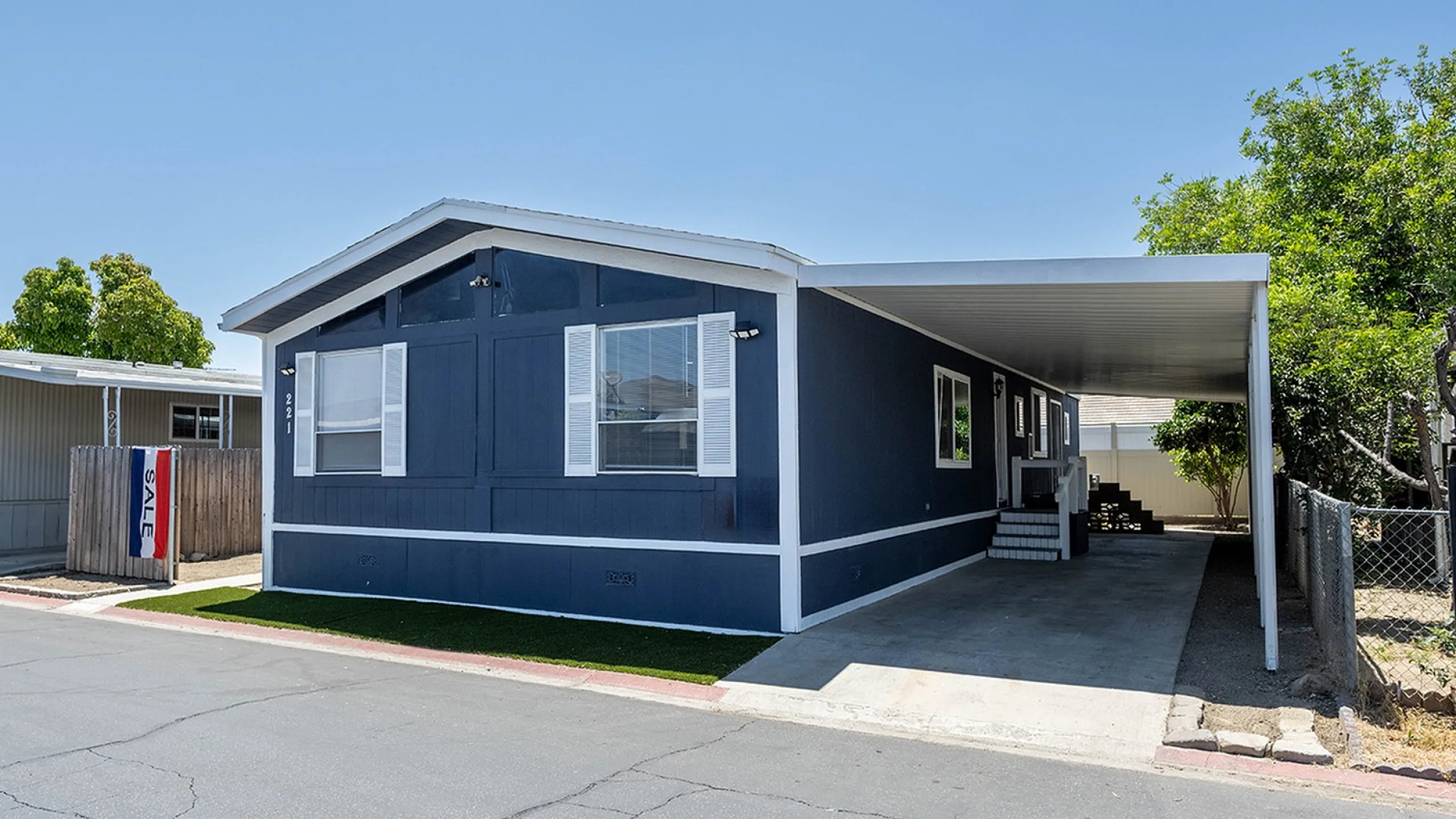 Charming mobile home with a sleek dark exterior, white trim, and a spacious covered carport under a clear blue sky.