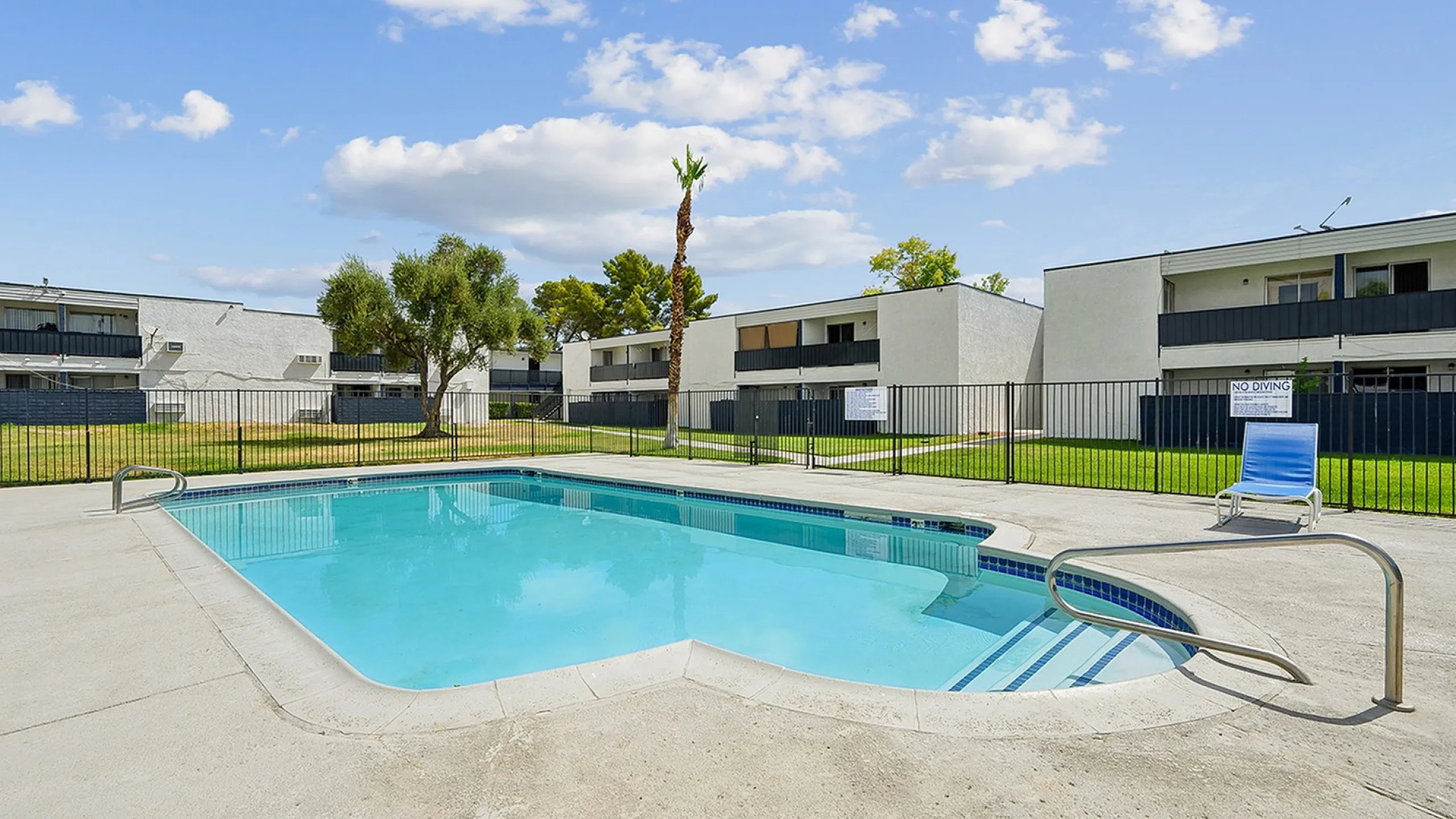 Inviting swimming pool area with modern lounge chair, surrounded by lush green landscape and contemporary apartment buildings under a sunny blue sky.