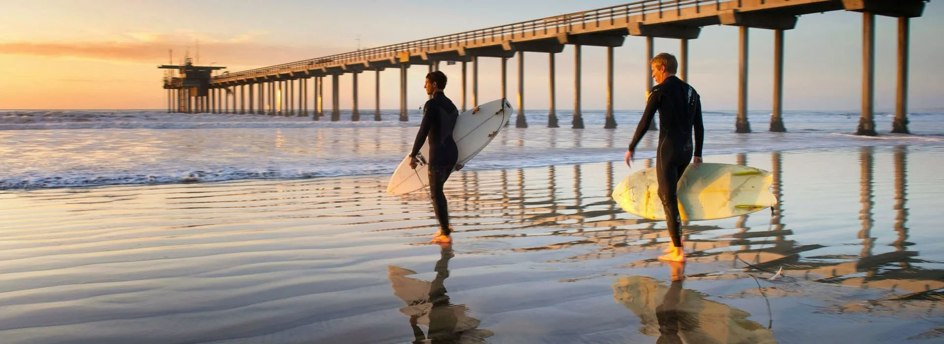 two people with surfboards on beach near a pier
