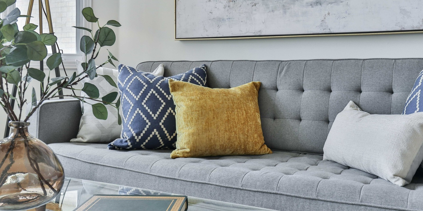 Grey couch with white, blue and gold throw pillows with glass coffee table and plants