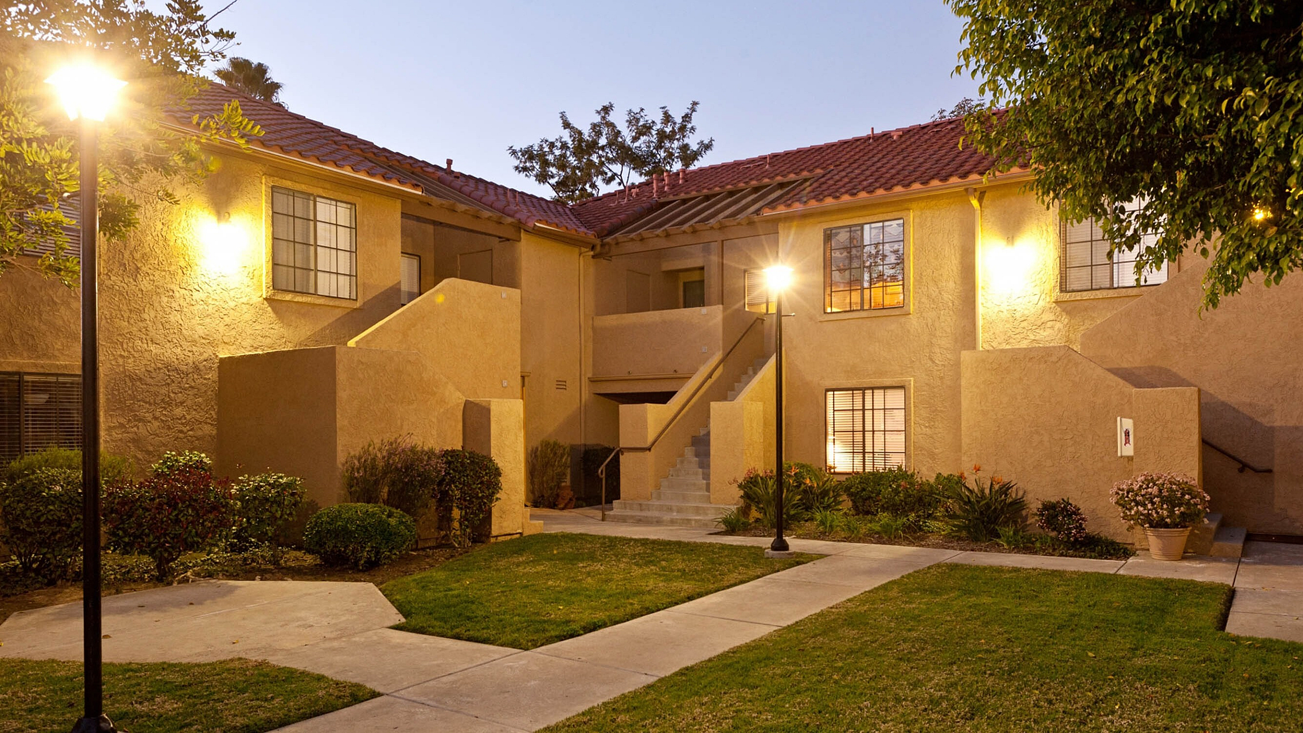 A wide concrete walkway leading through a manicured green lawn and professional landscaping toward the apartment entrances.