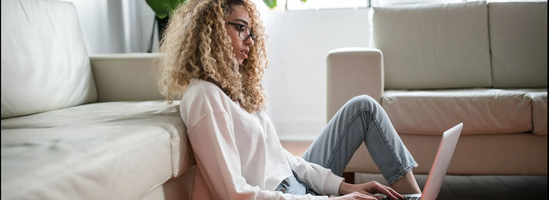 woman sitting on the floor, leaning against a white sofa while using a laptop