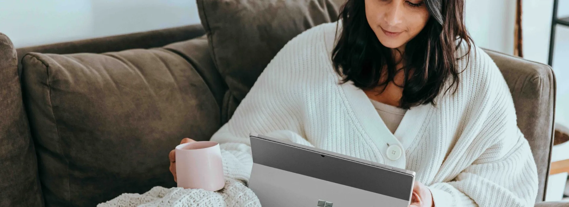 woman using a tablet while laying on a sofa drinking tea
