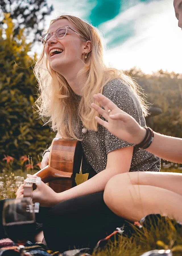 young woman playing the guitar and laughing while sitting on the grass next to another person