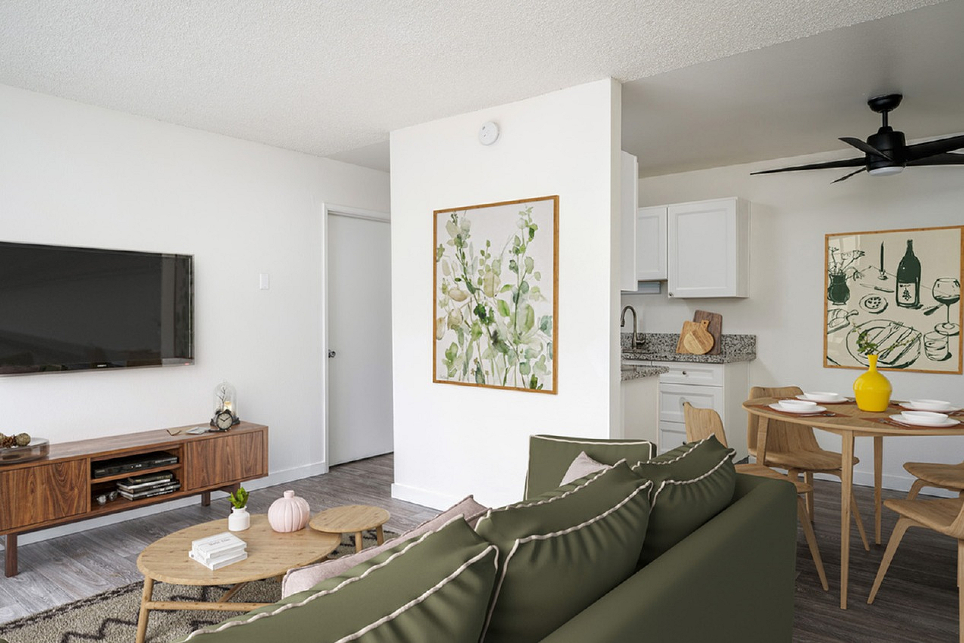 virtually staged living room with faux hardwood flooring and a green sofa with two coffee tables in front an entertainment console with a tv on the wall and a kitchen with a round dining table at boulder gardens