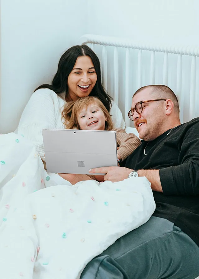 mom and dad cuddle with child in bed looking at a tablet