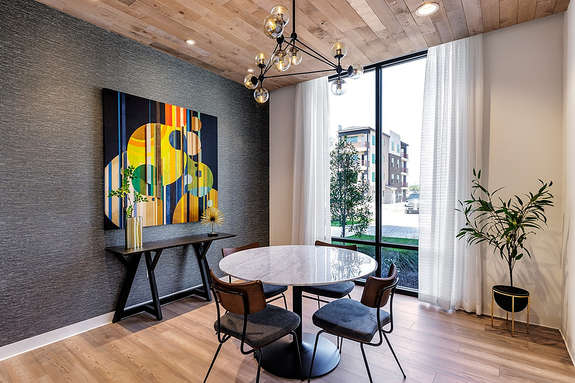 Contemporary dining area with a round marble table, modern chairs, a bold abstract painting, wooden ceiling, and large window.
