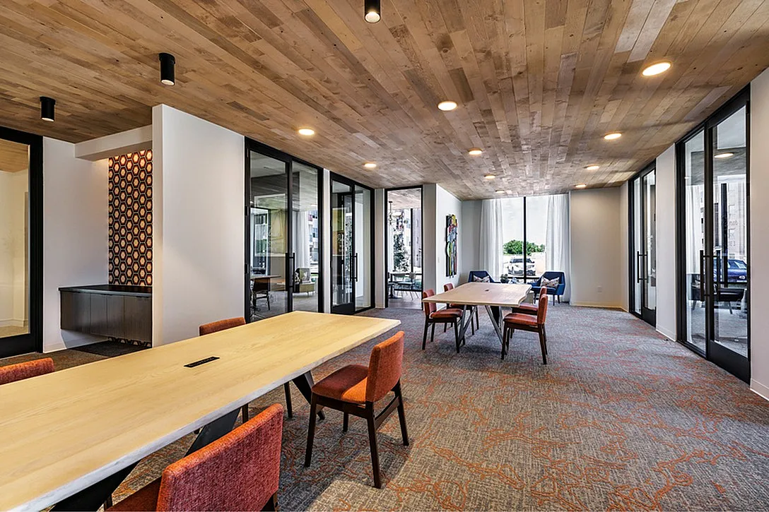 Stylish meeting lounge featuring modern wooden ceiling, long tables, vibrant chairs, and floor-to-ceiling windows.