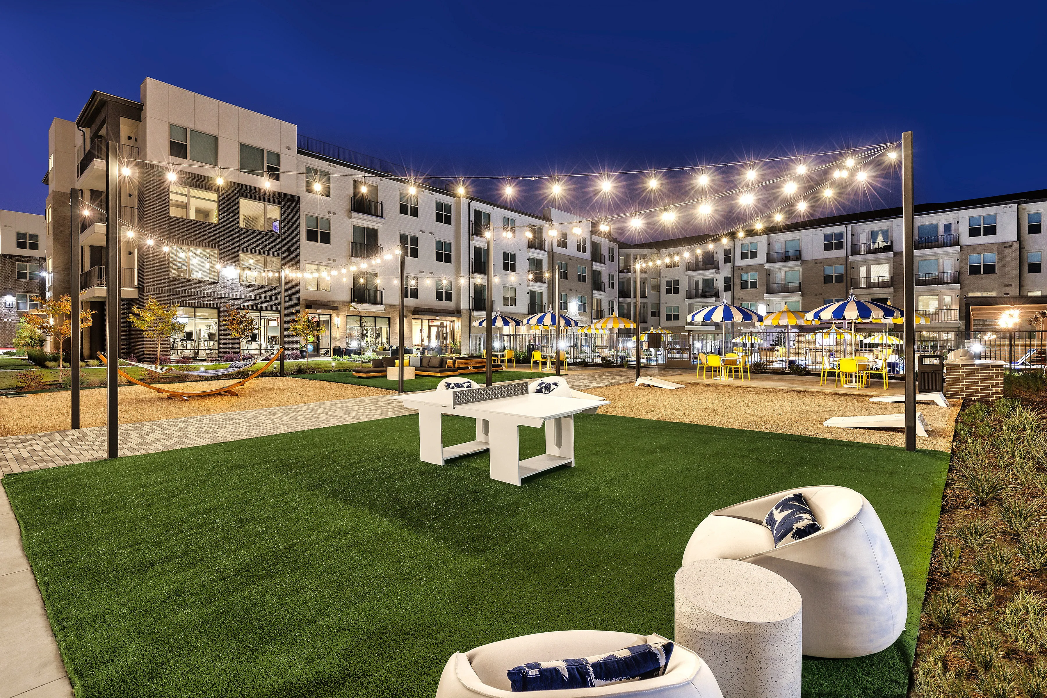 Illuminated courtyard with modern furniture, string lights, and elegant apartment building in the evening.