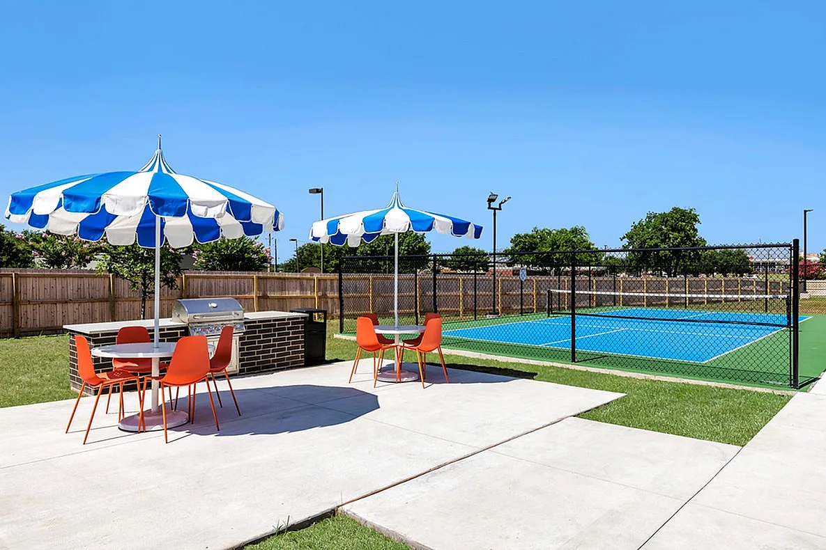 Outdoor area with blue and white umbrellas, orange chairs, and a tennis court.