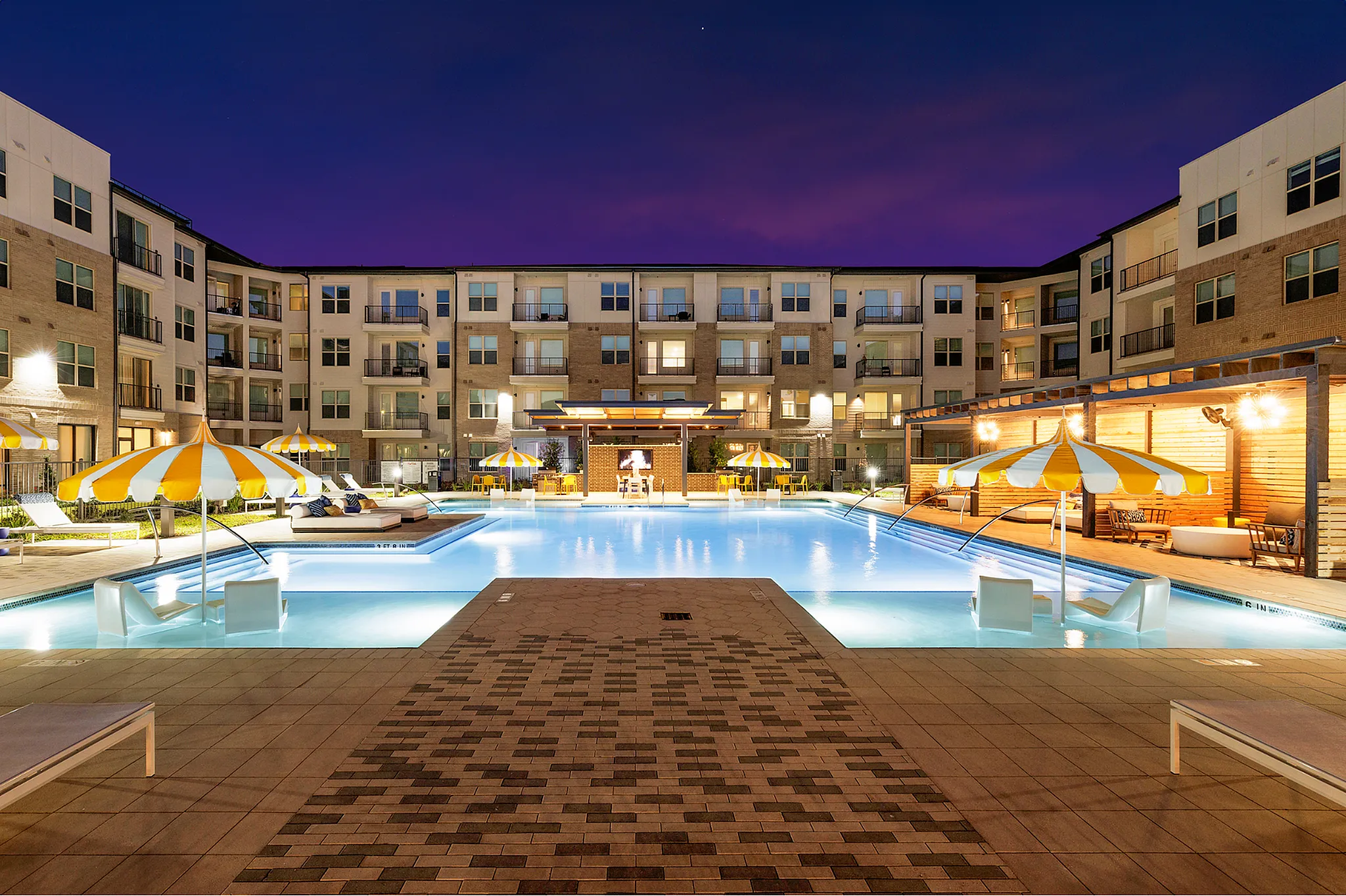 Modern apartment complex showcasing a well-lit pool area with stylish seating and umbrellas at night.