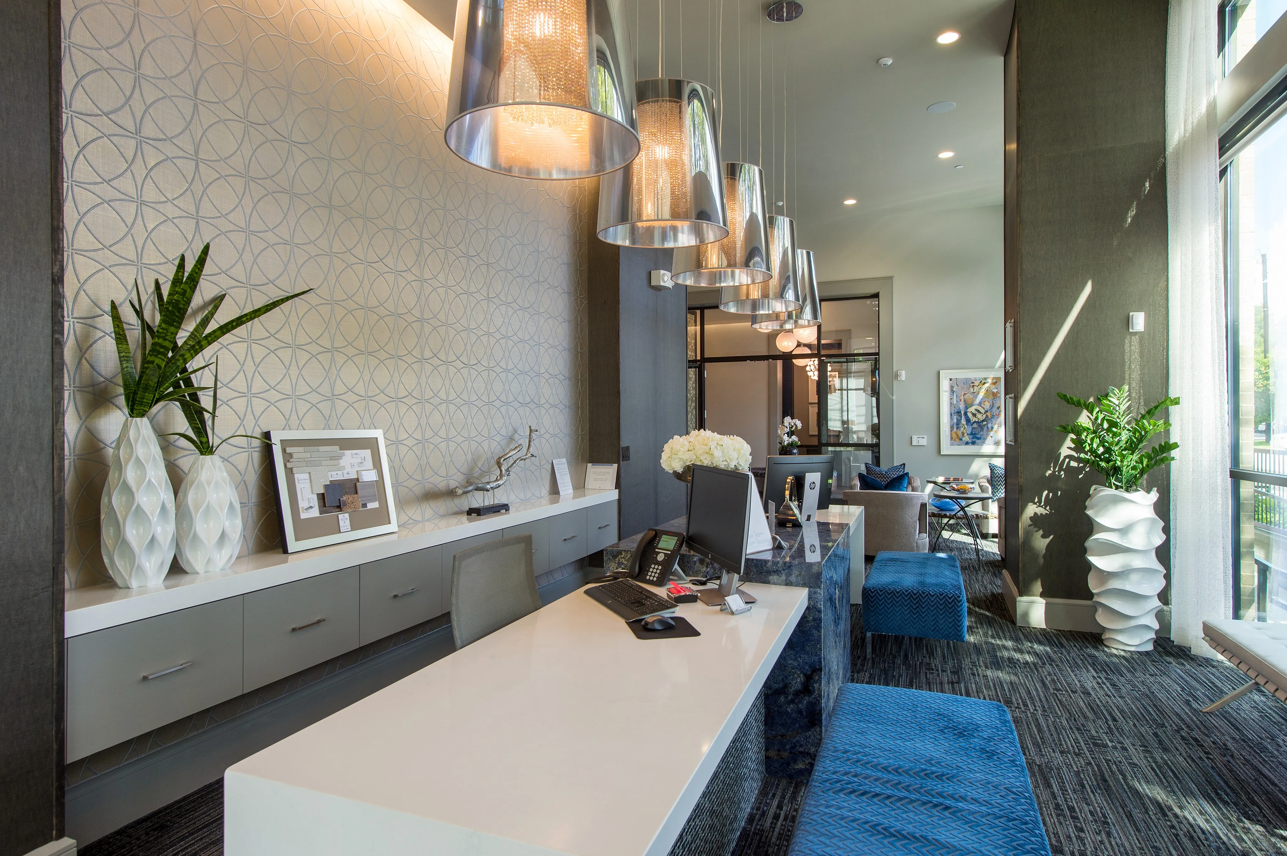 Luxurious and modern lobby with elegant decor, featuring stylish pendant lighting, a sleek white desk, vibrant blue seating, and lush greenery.