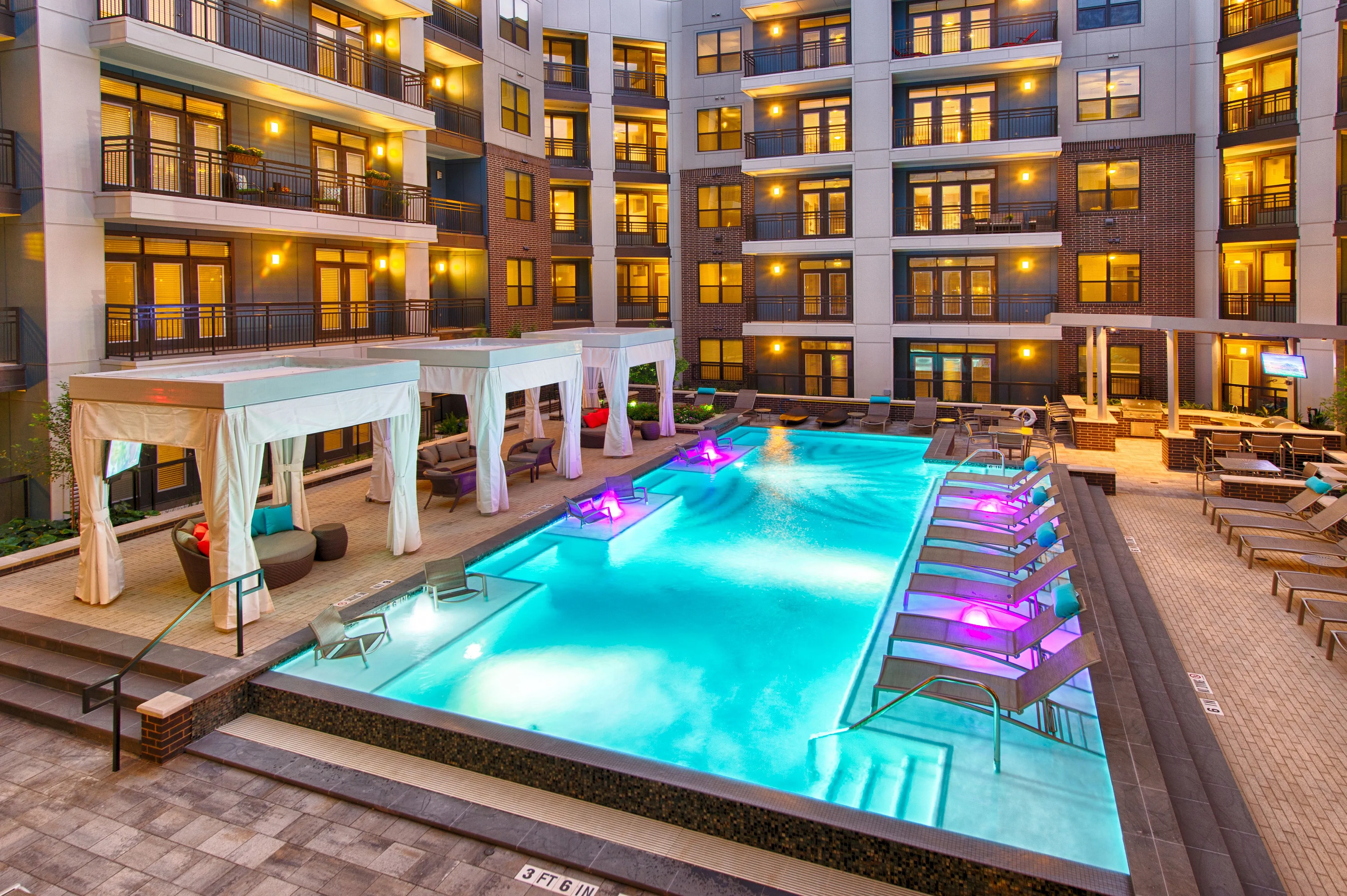 Luxurious apartment complex with a sparkling illuminated pool, elegant poolside cabanas, and modern balconies creating an inviting evening ambiance.