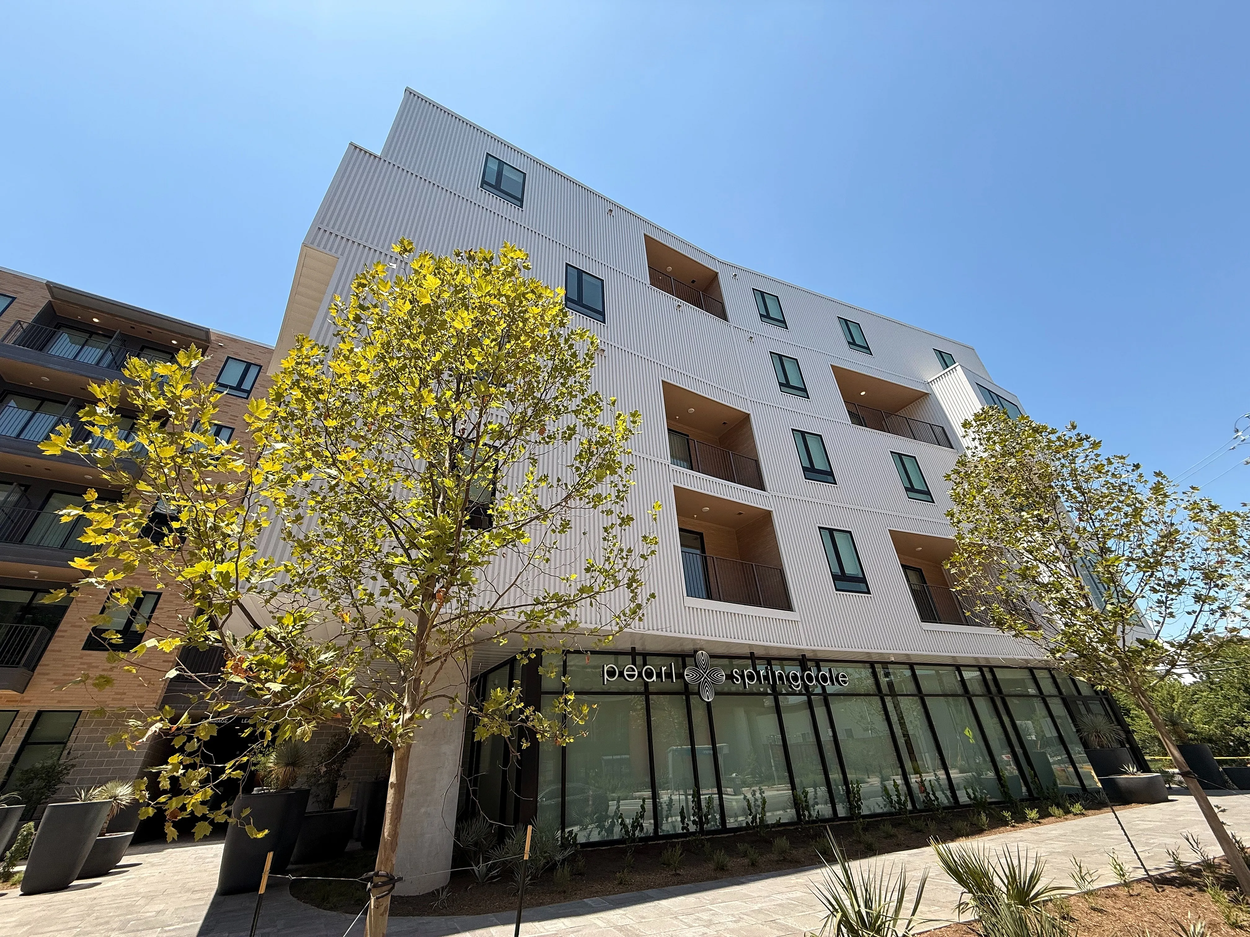 Modern apartment building with sleek design and elegant balconies, surrounded by lush trees under a clear blue sky.