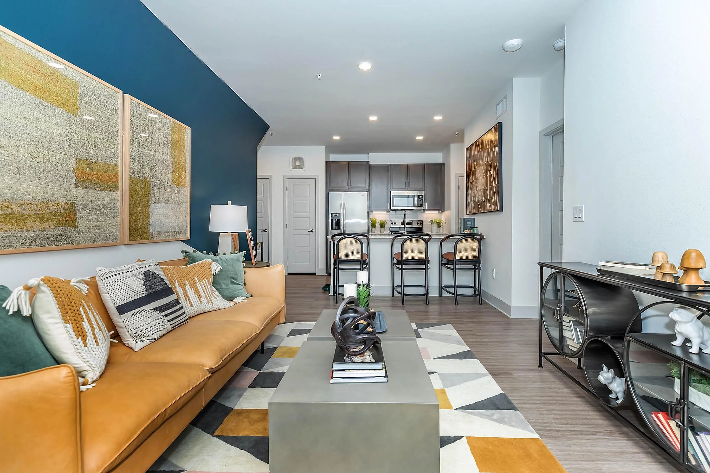 Modern living space with chic decor, featuring a stylish tan sofa with vibrant cushions, a sleek coffee table, and a cozy open-concept kitchen with bar seating.
