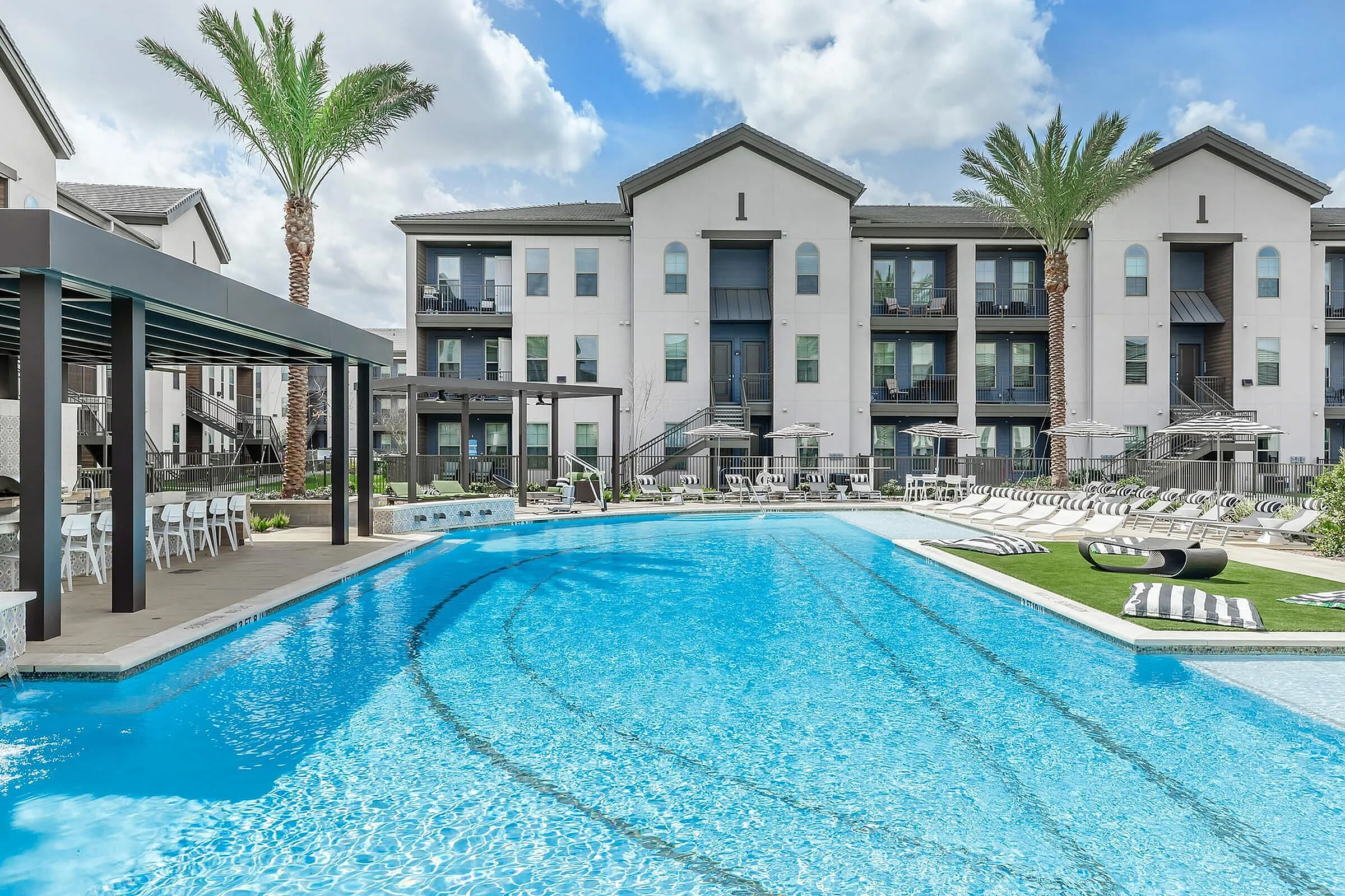 Luxurious apartment complex with modern architecture, featuring a refreshing swimming pool and stylish outdoor lounge area under sunny skies.