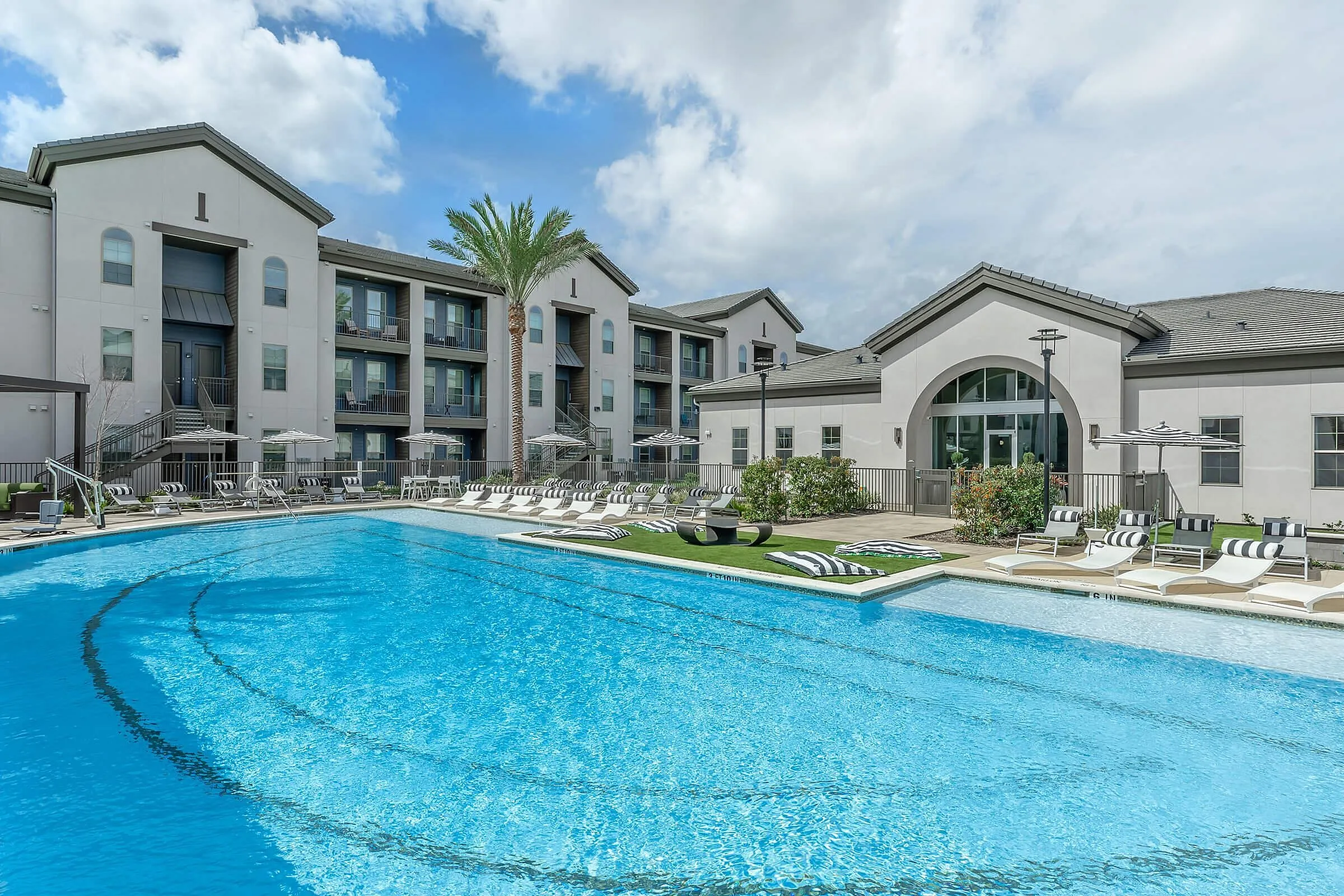 Luxurious apartment complex featuring a sparkling outdoor pool, modern architecture, and inviting lounge chairs set against a clear blue sky.