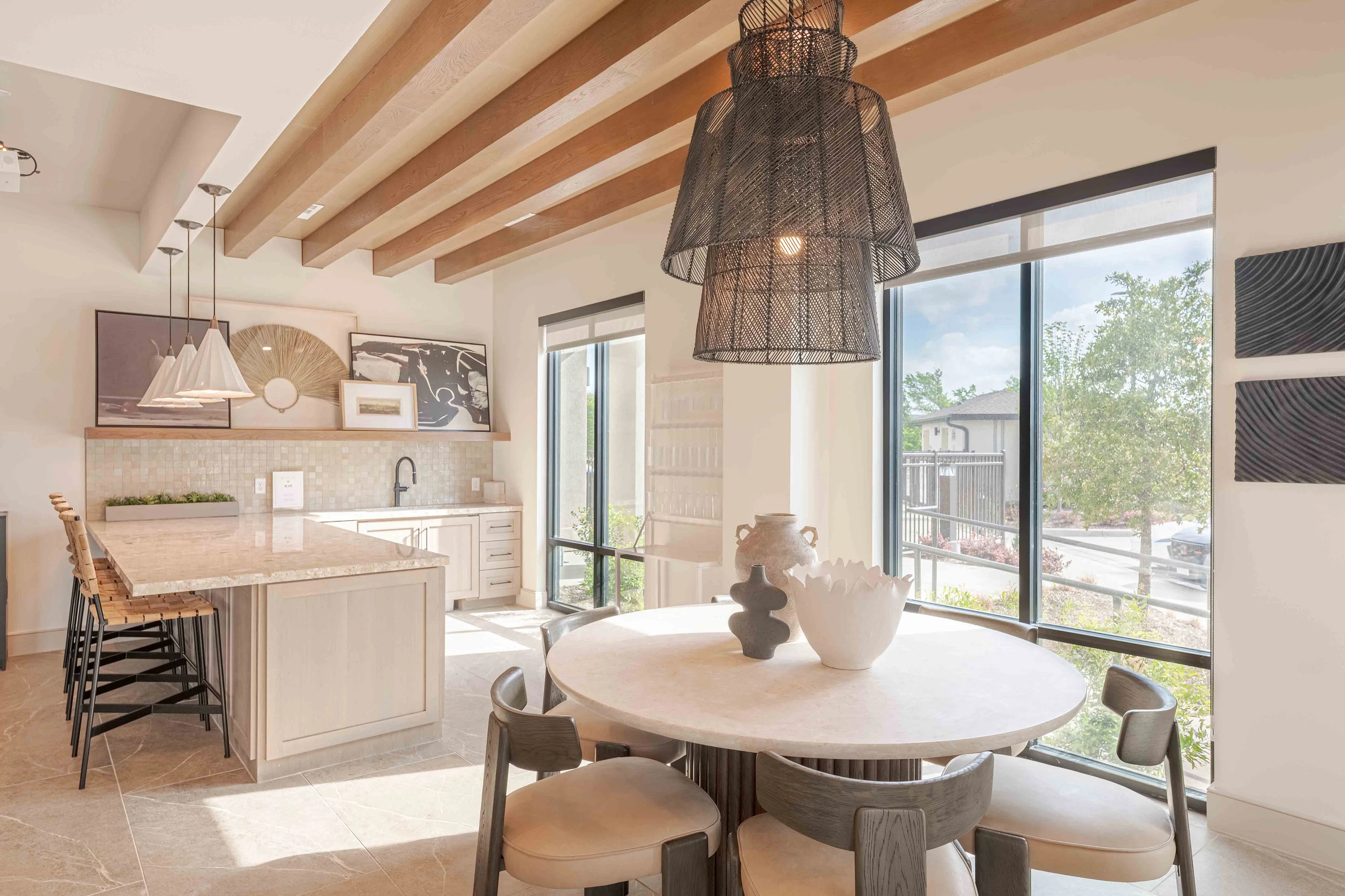 Chic dining area with large windows and wooden beams, featuring a modern kitchen with pendant lighting and decorative accents.