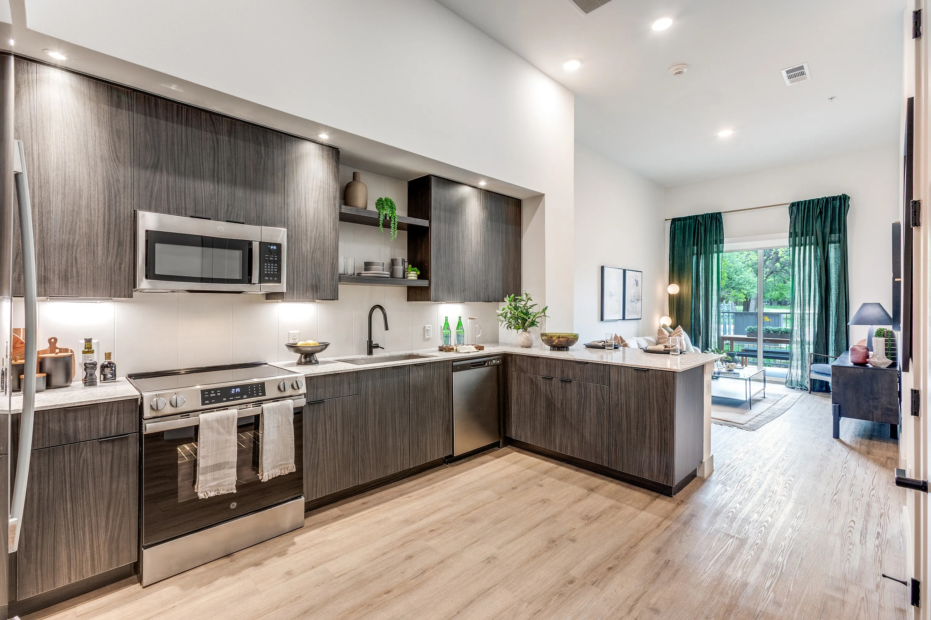 Contemporary kitchen and living room with sleek dark cabinetry, stainless steel appliances, and natural light from large windows.