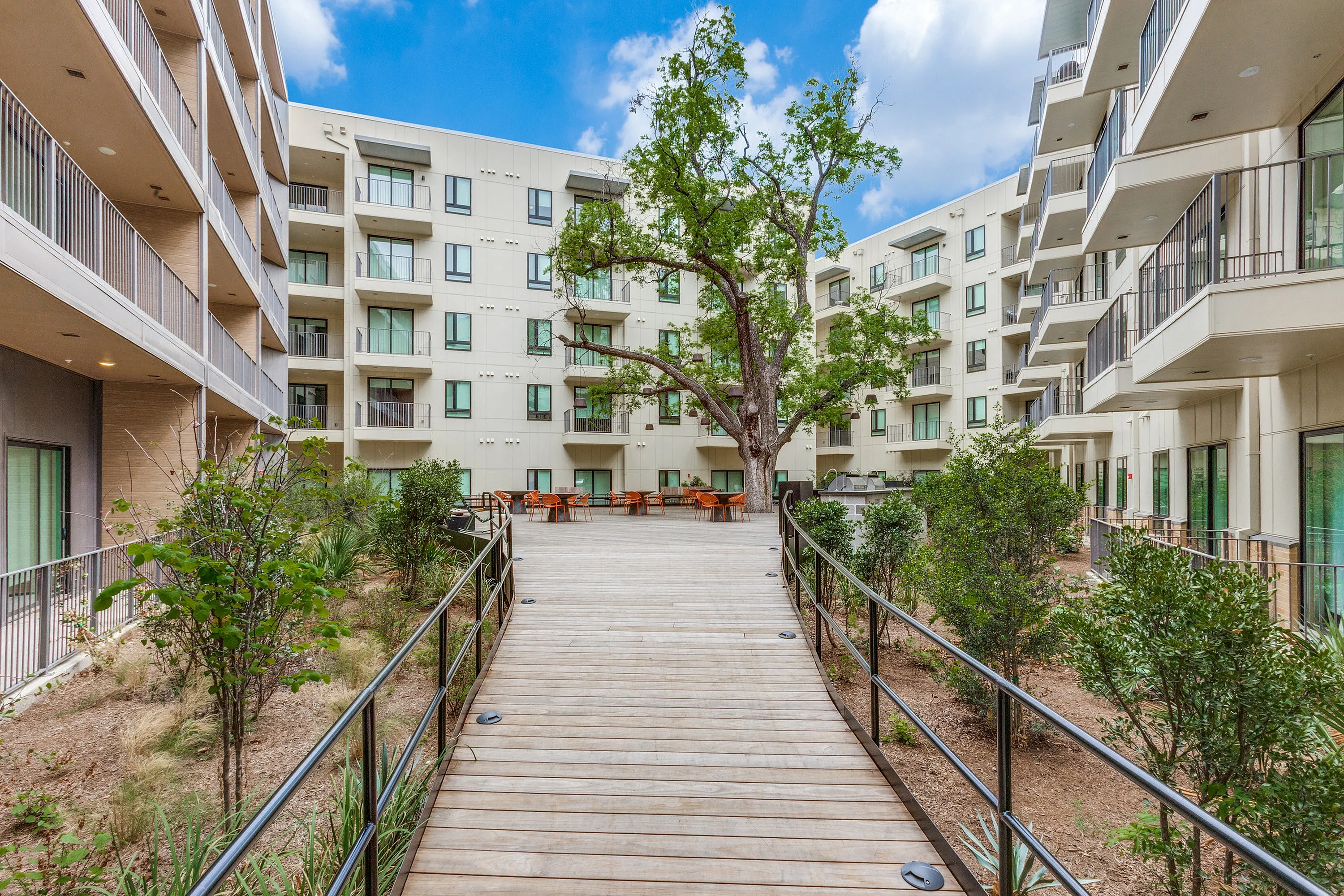 Modern apartment complex featuring lush greenery and a serene wooden walkway leading to outdoor seating beneath a towering tree.