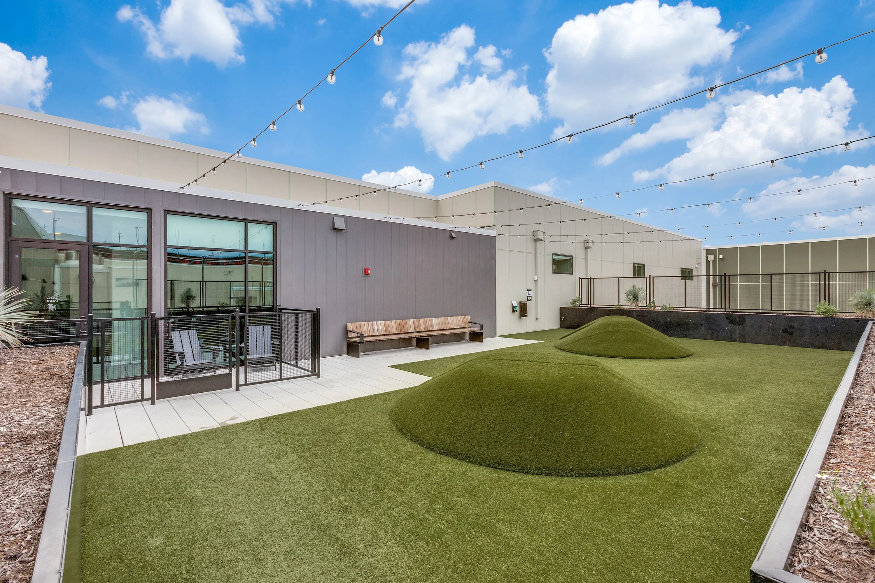 Modern rooftop terrace with lush green artificial turf, cozy seating area, and string lights under a vibrant blue sky.