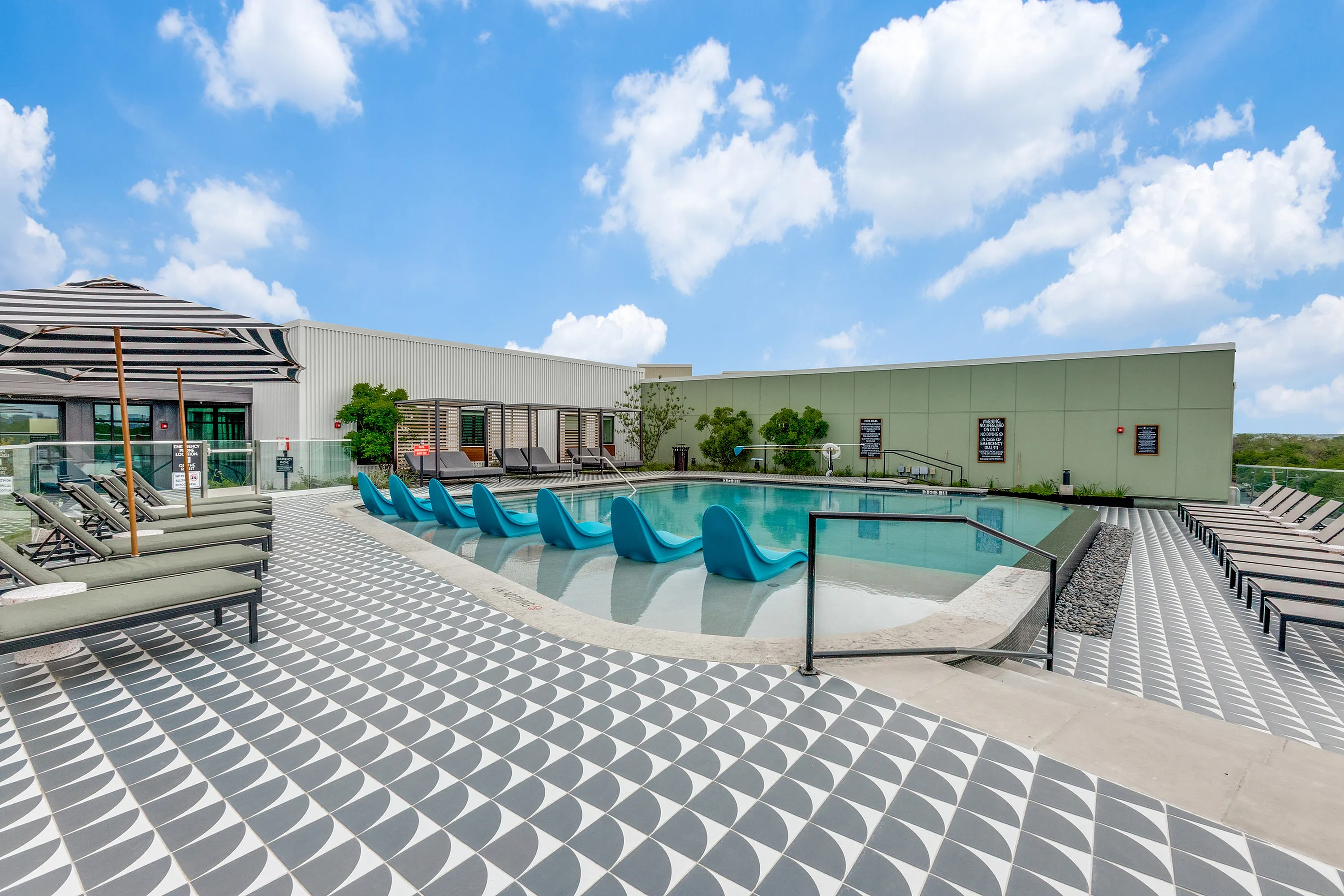 Luxurious outdoor pool area with modern blue lounge chairs, chic geometric tiling, and shaded cabanas under a vibrant sky.