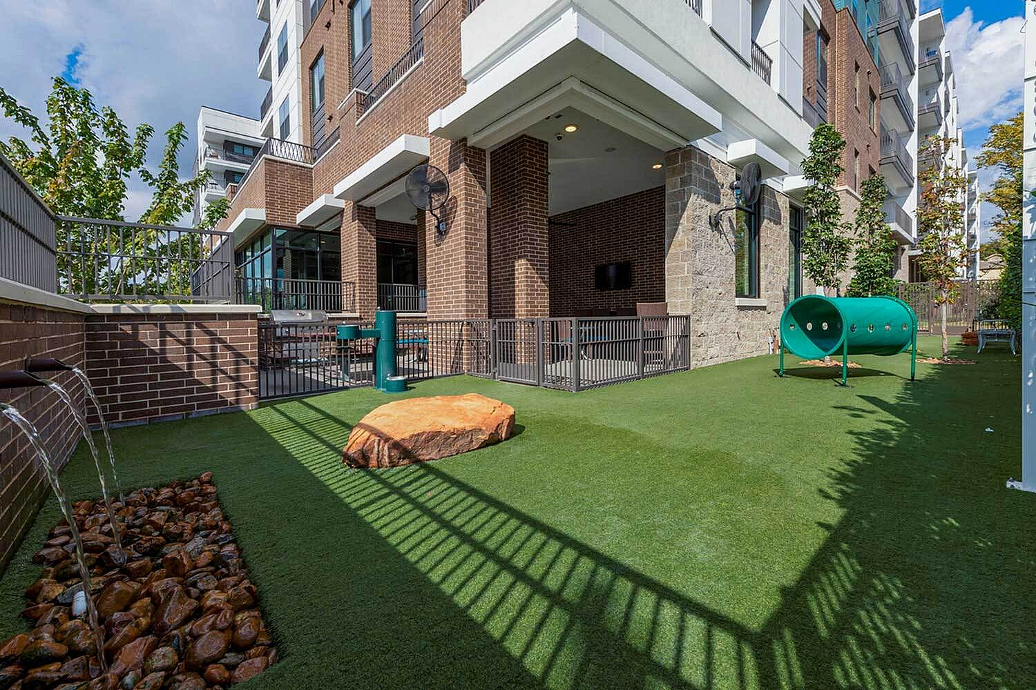 Inviting outdoor space showcasing a modern apartment complex with a green grassy area, playful dog tunnel, and elegant water feature.