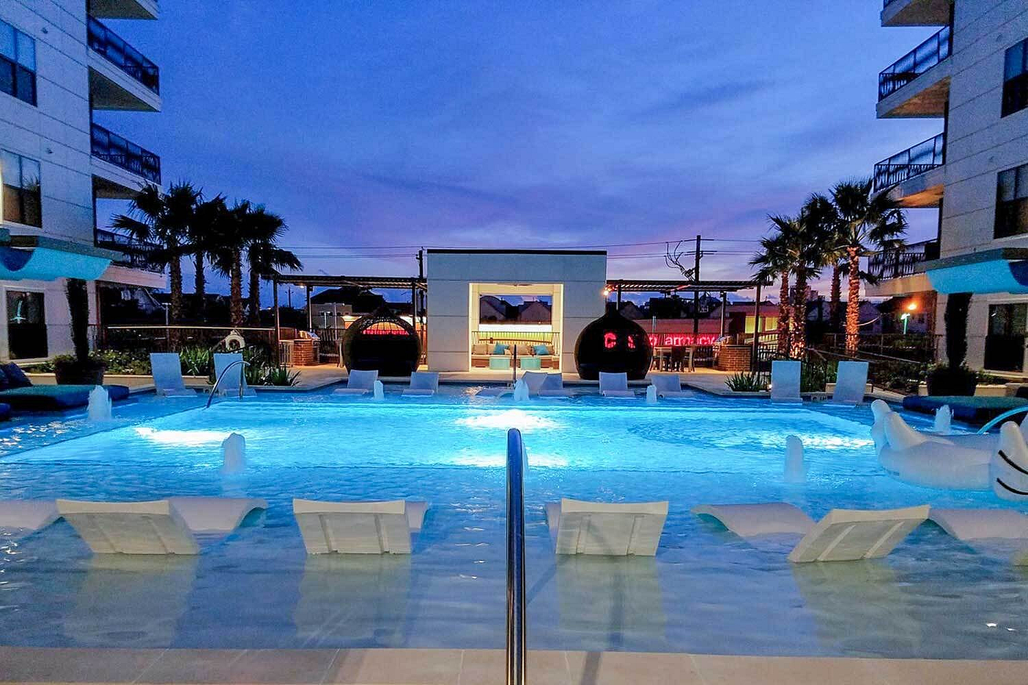 Luxurious outdoor pool area at dusk, featuring chic lounge chairs submerged in crystal-clear water, surrounded by modern architecture and swaying palm trees.