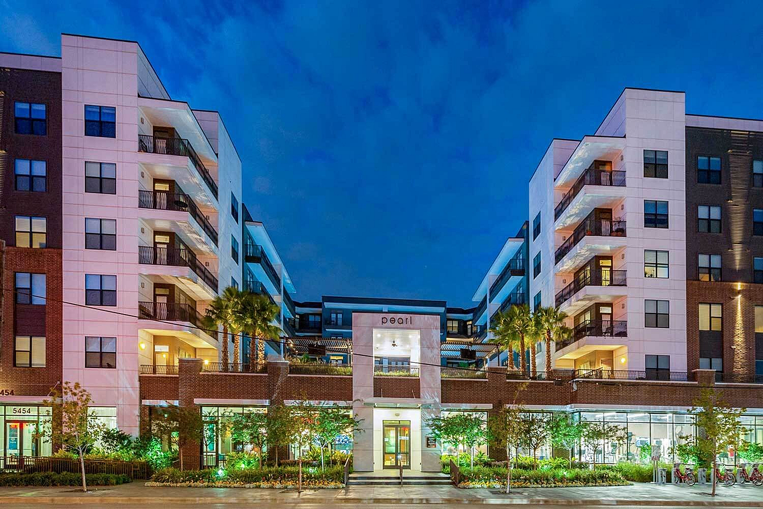 Modern urban apartment complex featuring stylish balconies, lush landscaping, and vibrant lighting against a twilight sky.