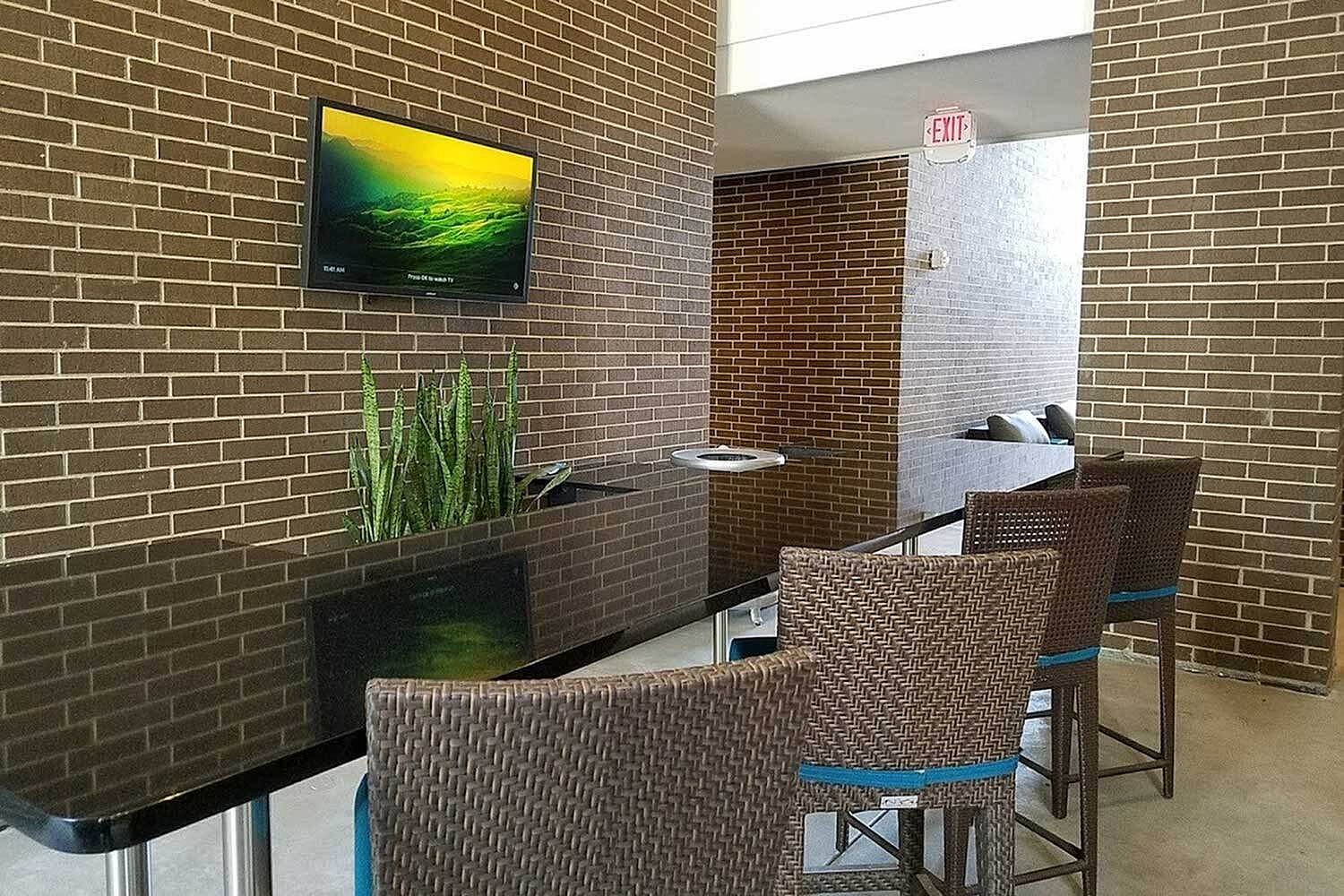 Sophisticated lounge area featuring modern wicker chairs, a sleek glass table, vibrant wall art, and lush greenery against an elegant brick backdrop.