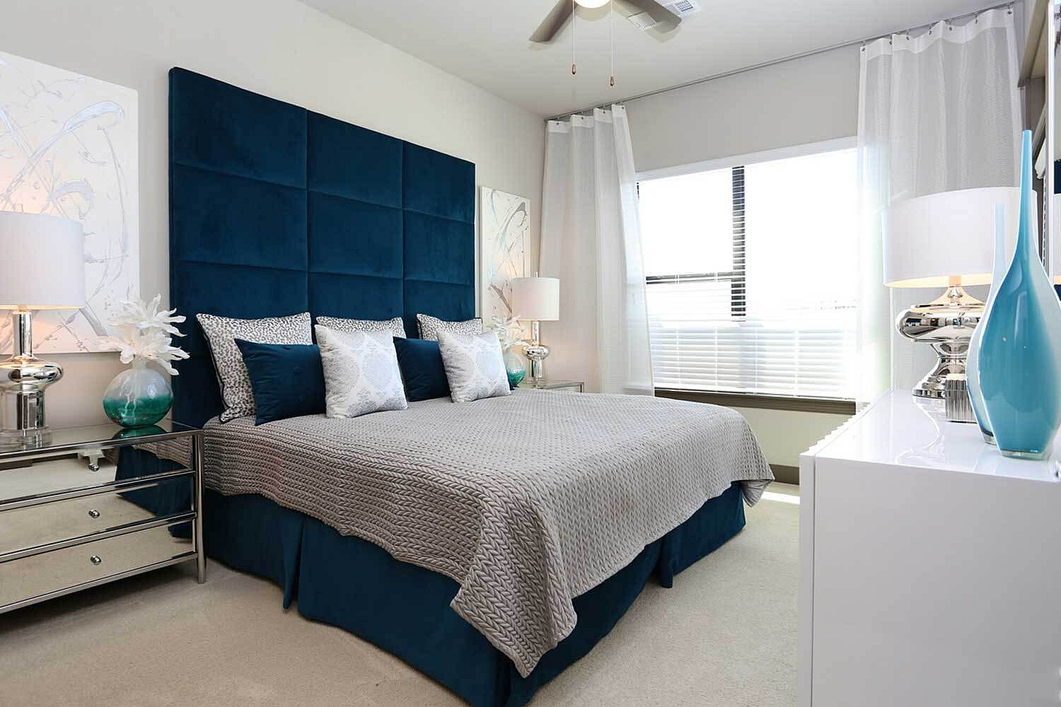 Luxurious bedroom with plush blue headboard, elegant decor, and large sunlit window.