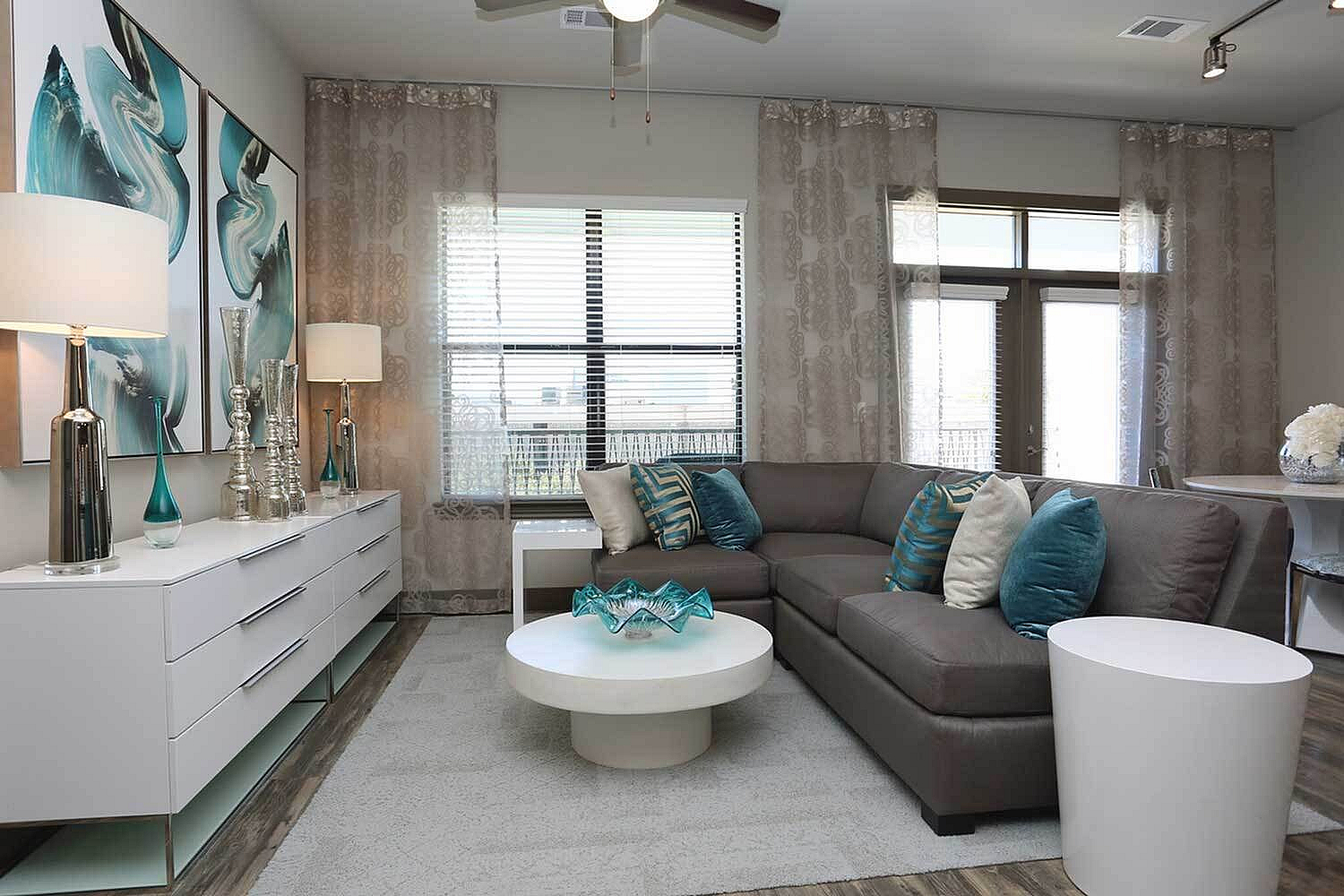 Modern, elegantly decorated living room featuring a plush grey sectional couch, chic white coffee table with accent artwork, and abundant natural light through large windows.